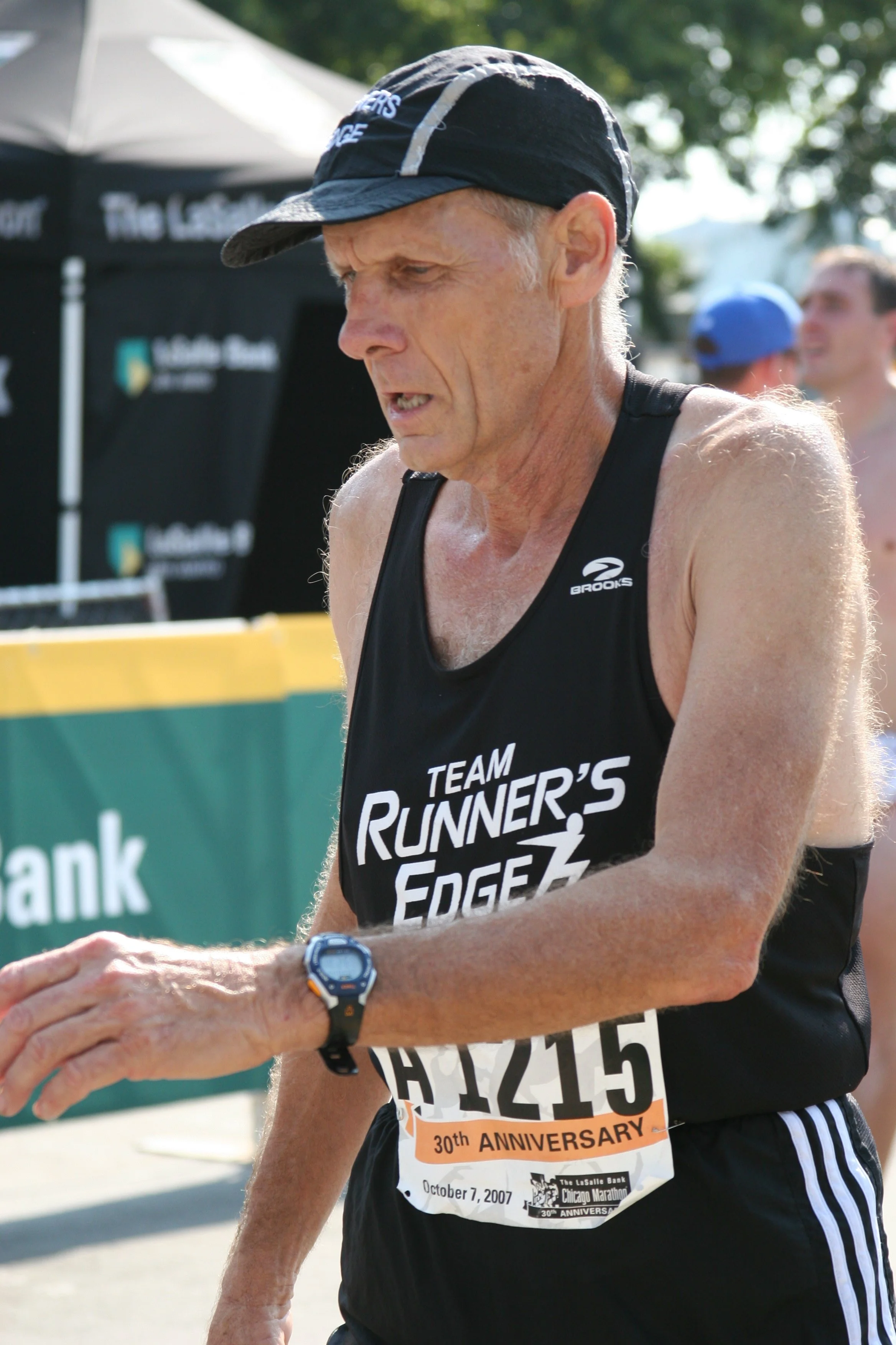A male runner in athletic gear participating in a marathon, wearing a black tank top with the words 'TEAM RUNNER'S EDGEZ' and a race bib number 1645. He appears to be sweaty and focused, with a black cap and a wristwatch. The background shows a green