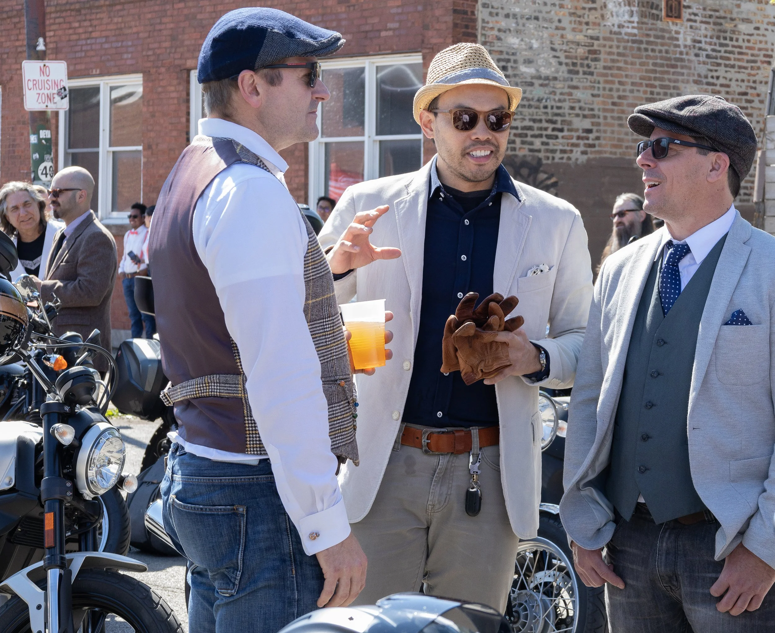 Three men in vintage style clothing talking outdoors near motorcycles, with a brick building in the background.