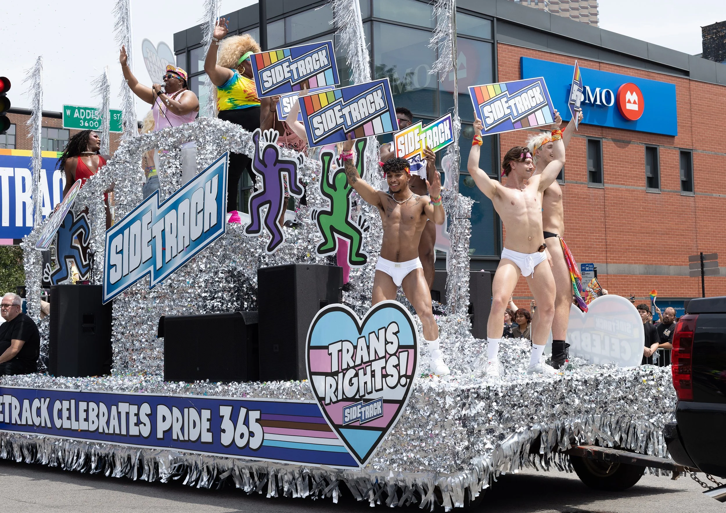 A parade float celebrating Pride, decorated with silver fringe and logos for 'Side-Track'. Several people, mostly shirtless, are dancing and holding signs that say 'Side Track'. The float is on a street with a brick building and an 'AMO' sign in the 