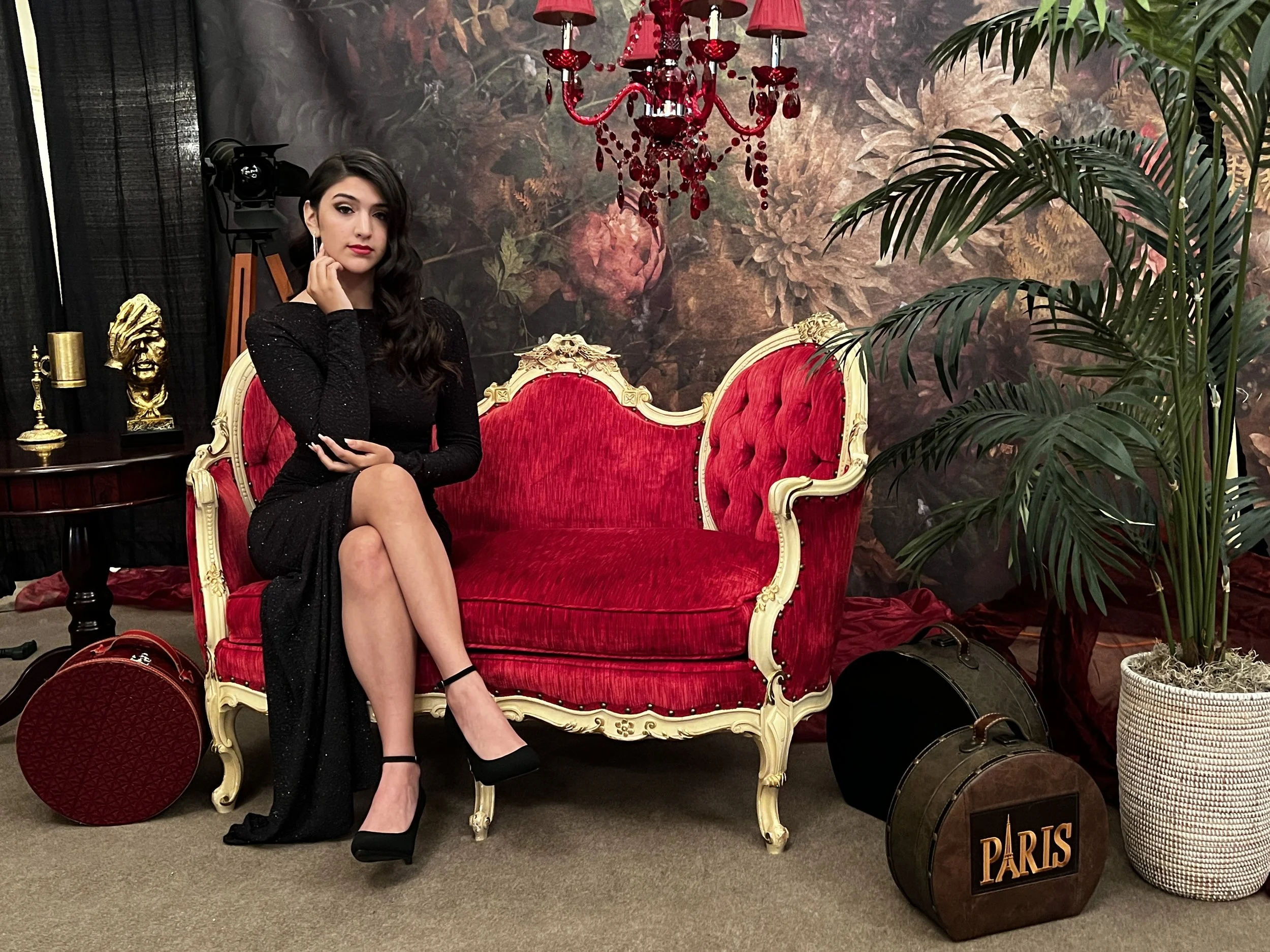 A woman with dark, wavy hair, wearing a black, long-sleeve, knee-length dress and black high heels, sitting on a red ornate vintage sofa with gold accents. She has her legs crossed and is resting her chin on her left hand, looking directly at the cam
