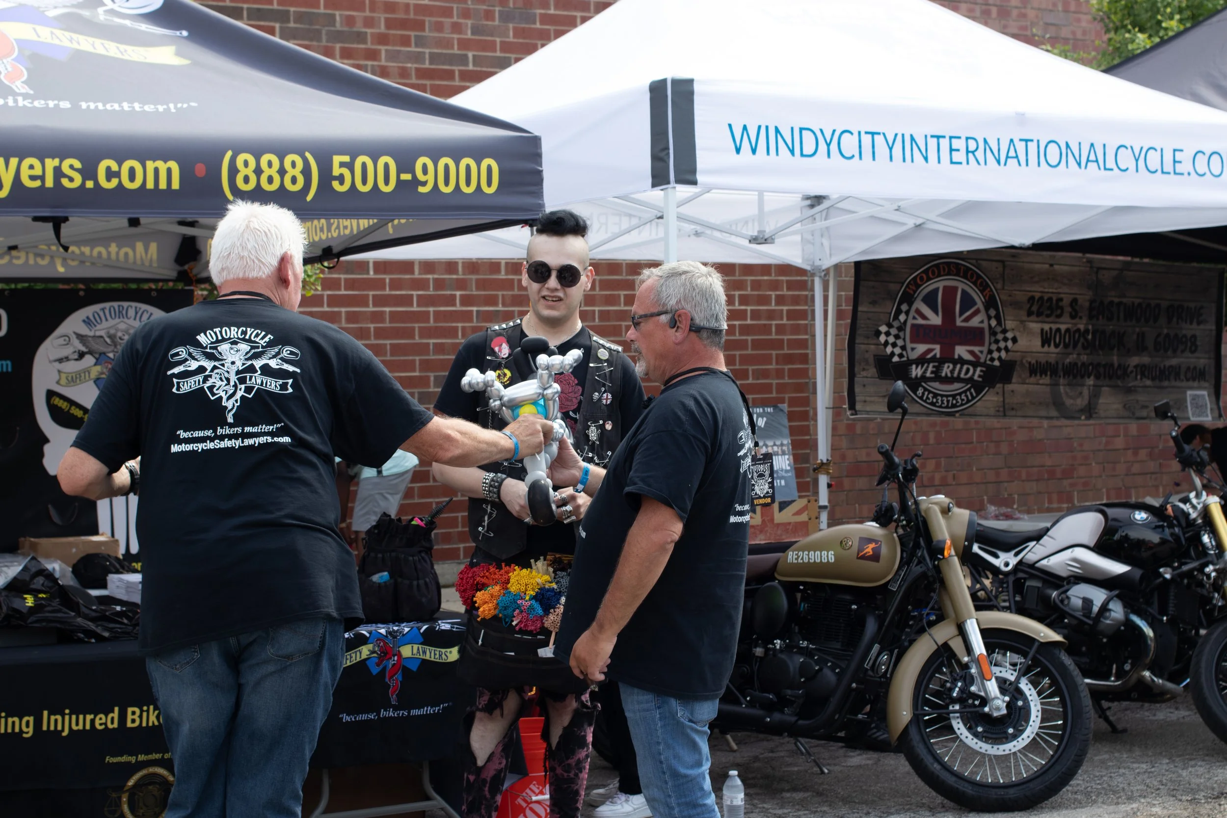 Three men at an outdoor booth with motorcycles and banners, engaging in a conversation. The booth has a blue and white canopy with text and a website address, and there is a bouquet of colorful artificial flowers on the table.