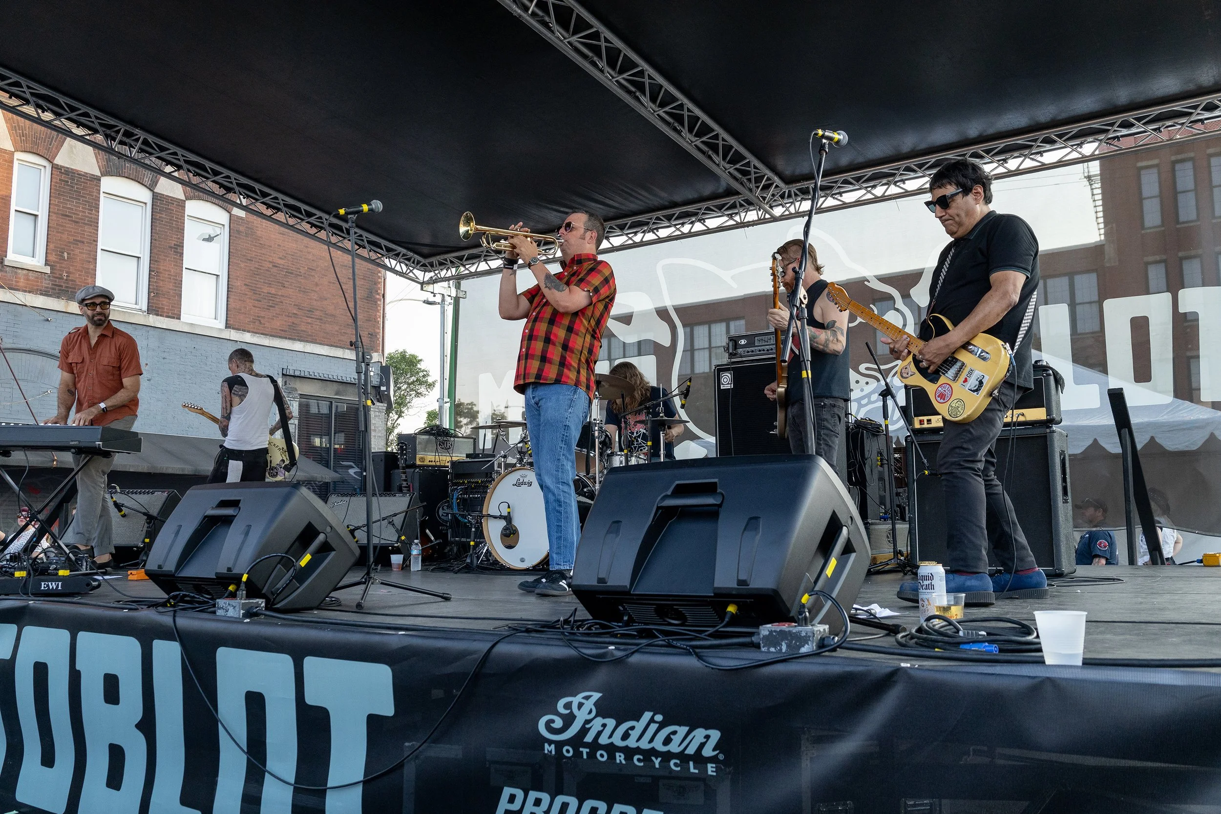 A live band performs on an outdoor stage, including a trumpet player, a guitarist, a keyboardist, and a bassist. The stage is covered by a black canopy and features various music equipment, with brick buildings visible in the background.