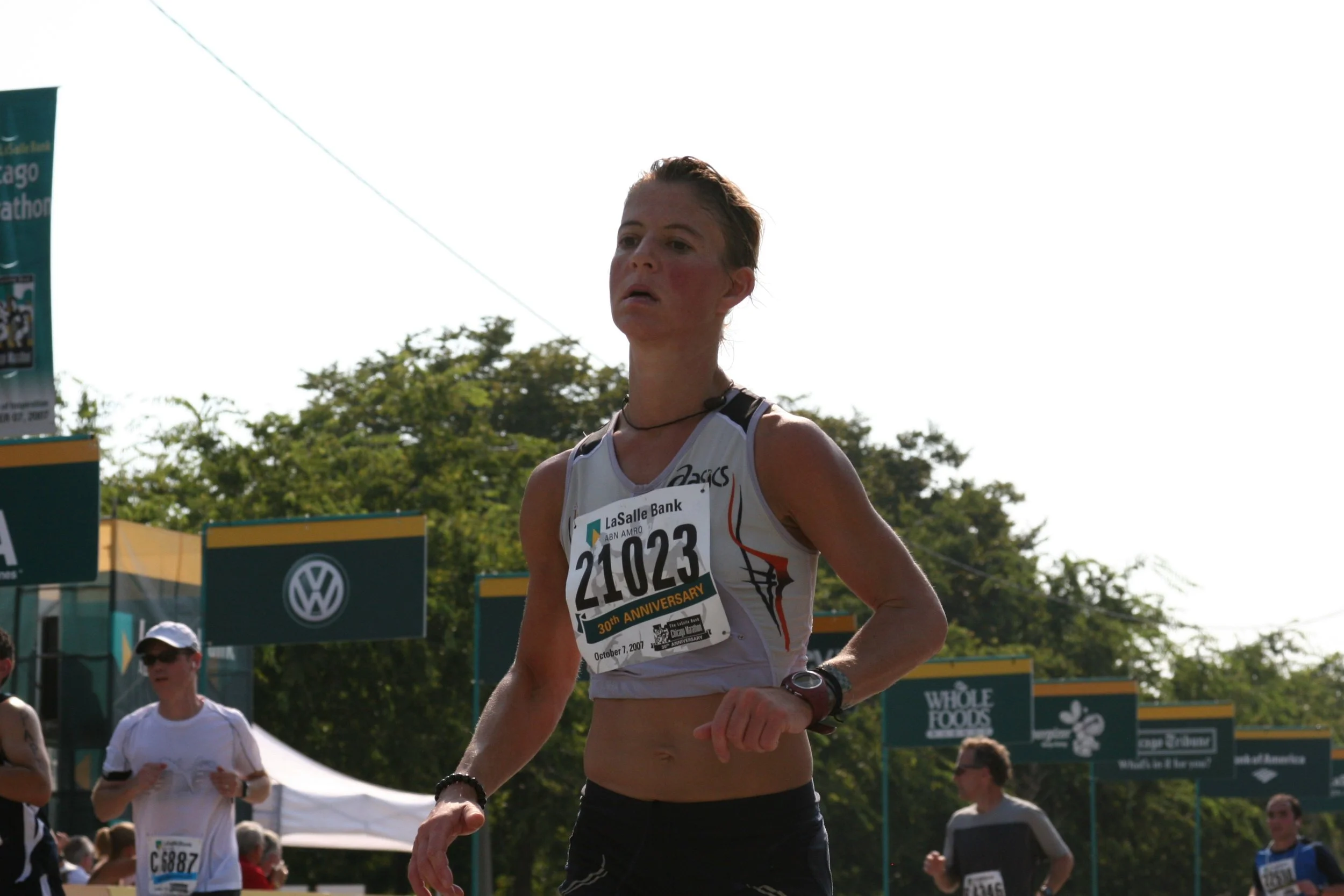 A female marathon runner with bib number 21023 participating in a race. She has a focused expression, wearing a sleeveless athletic top, and appears to be nearing the finish line. Other runners and race banners are visible in the background.
