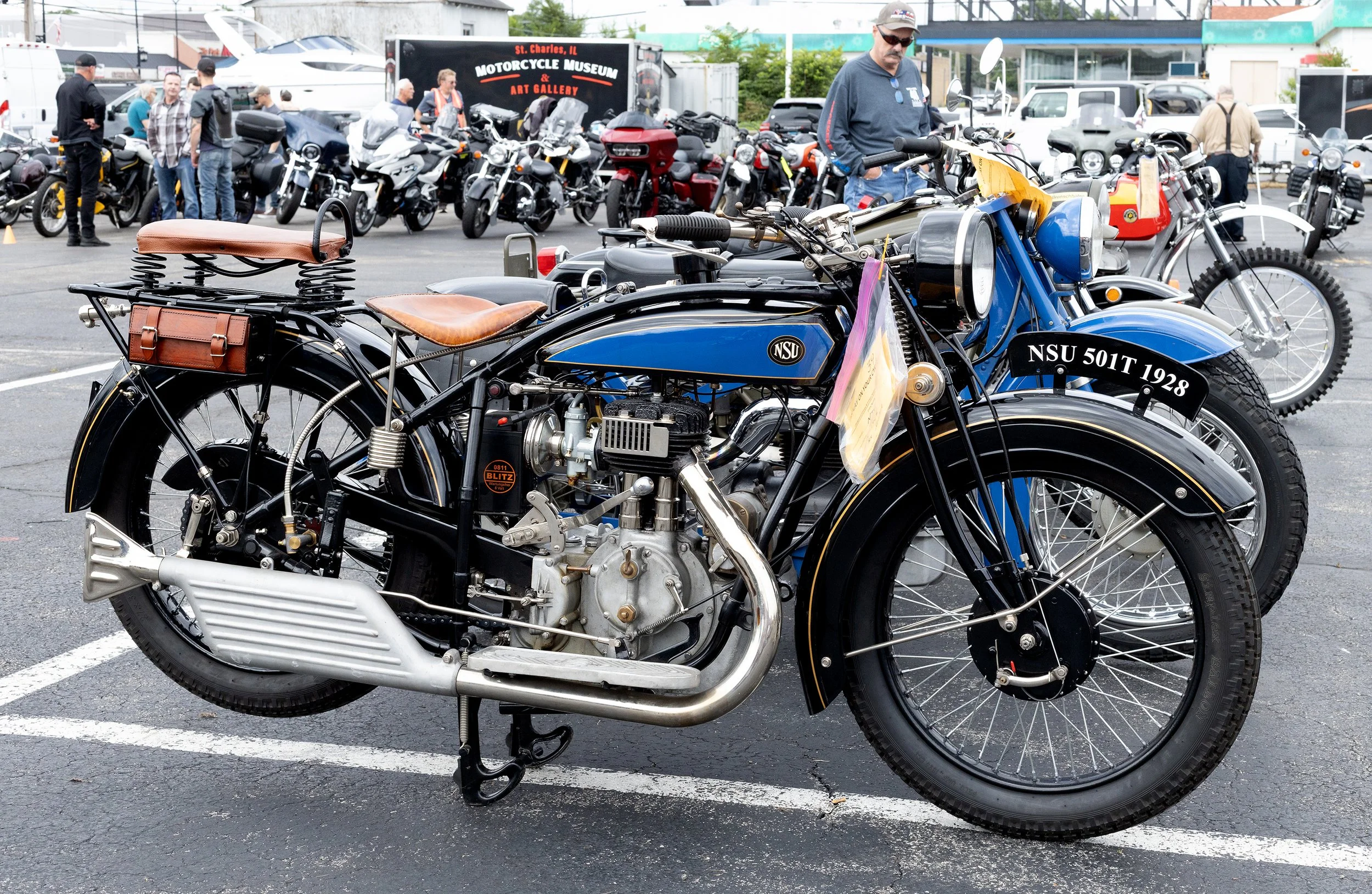Vintage motorcycle with leather saddle and tool bag parked among modern motorcycles at a motorcycle exhibition, with people viewing bikes in background.
