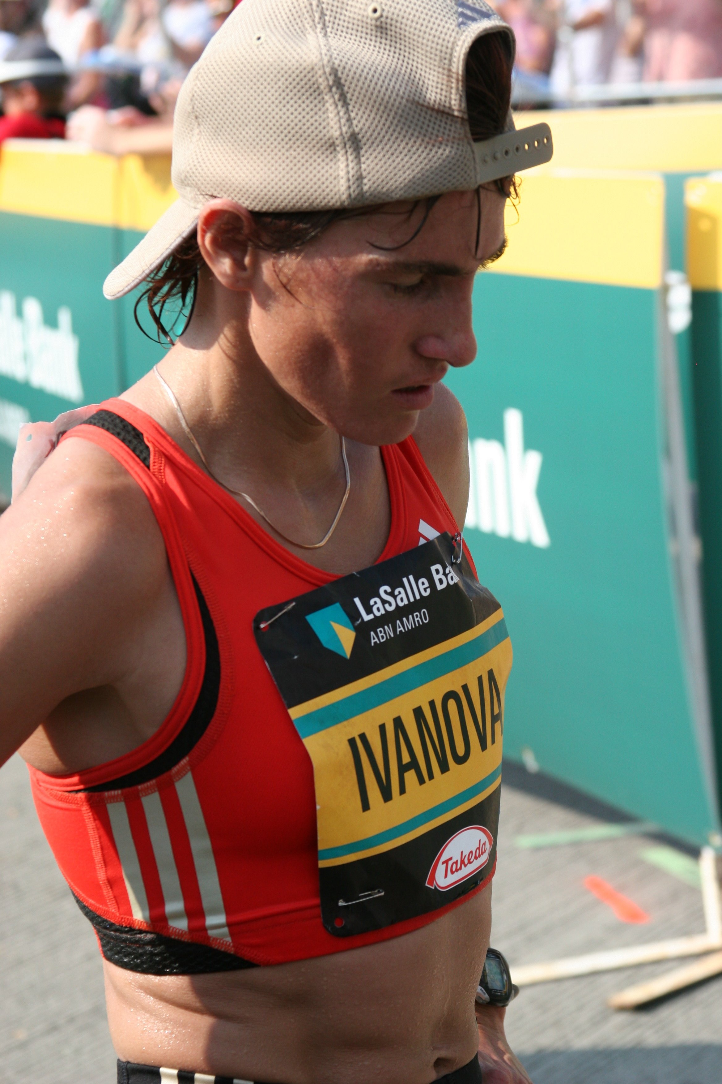 Close-up of a female marathon runner with wet hair wearing a beige cap backward, a red running tank top, and a yellow race bib with the name 'IVANOVA' and sponsor logos, during a race event.
