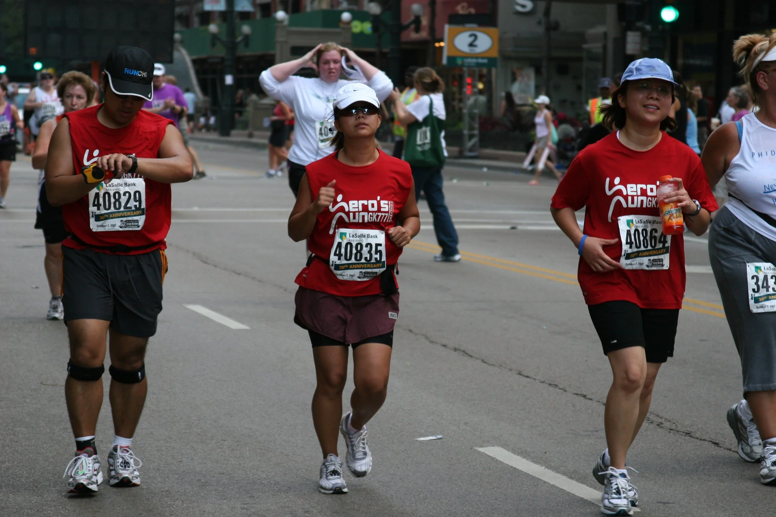 People participating in a marathon race, running on a city street with spectators in the background. Some runners are wearing red shirts and sunglasses, and one is holding a water bottle. The scene is set during daytime.