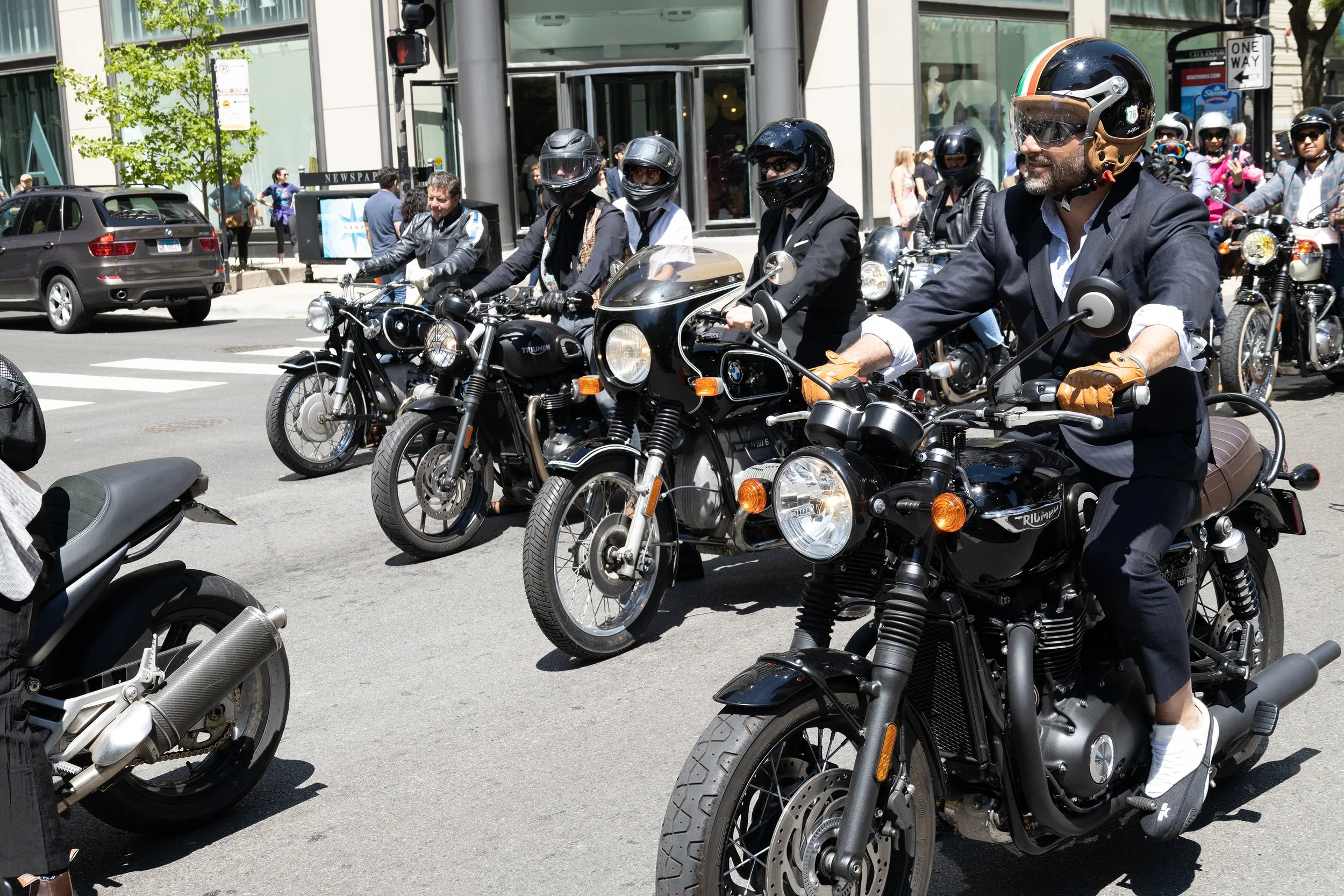 A group of motorcyclists on city street, wearing helmets and riding bikes, with pedestrians and buildings in the background.