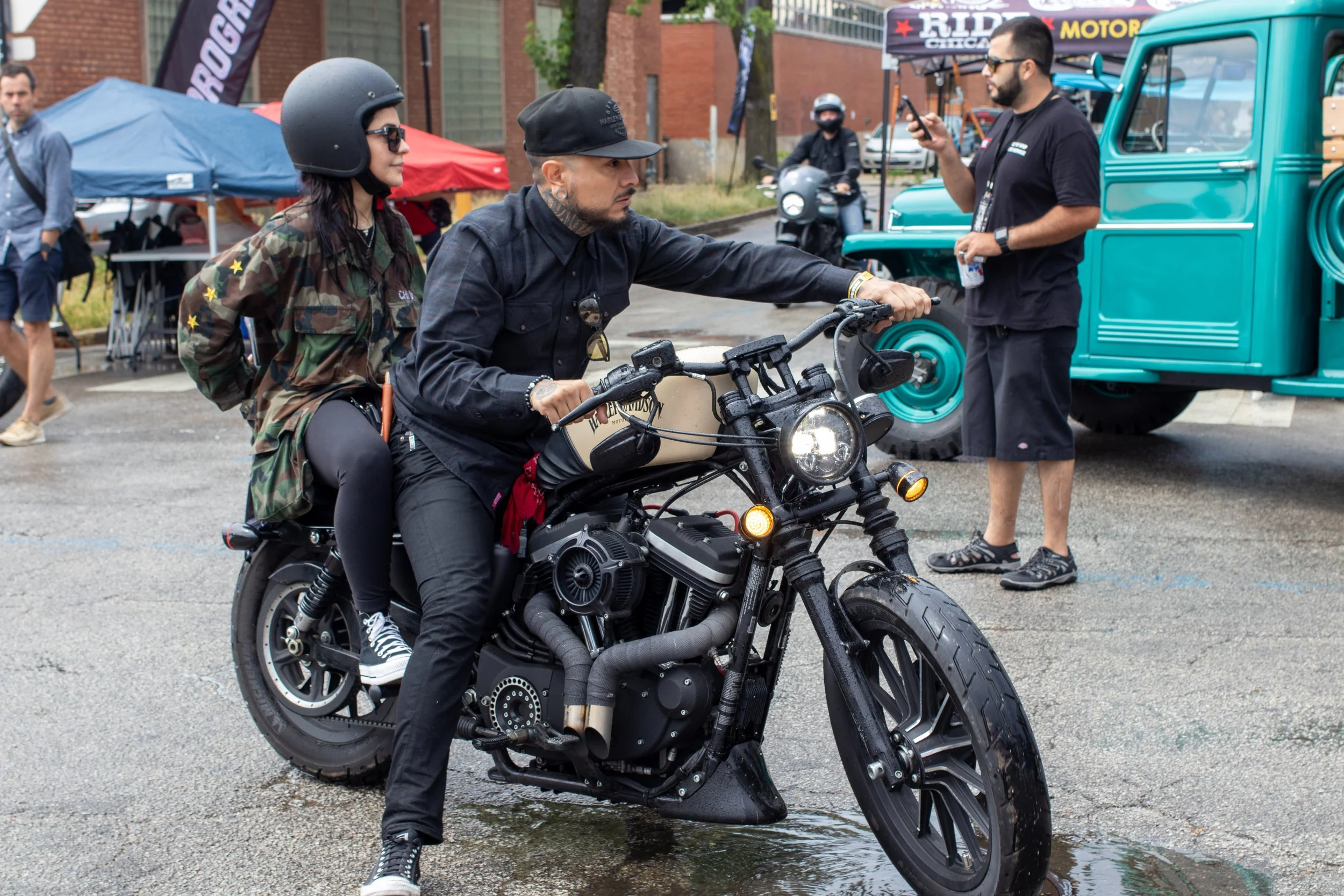 A woman wearing a helmet and sunglasses riding a black motorcycle with a man in black clothing, with tattoos, sitting behind her during an outdoor event. People are in the background, some wearing masks, with tents and a teal vehicle visible.