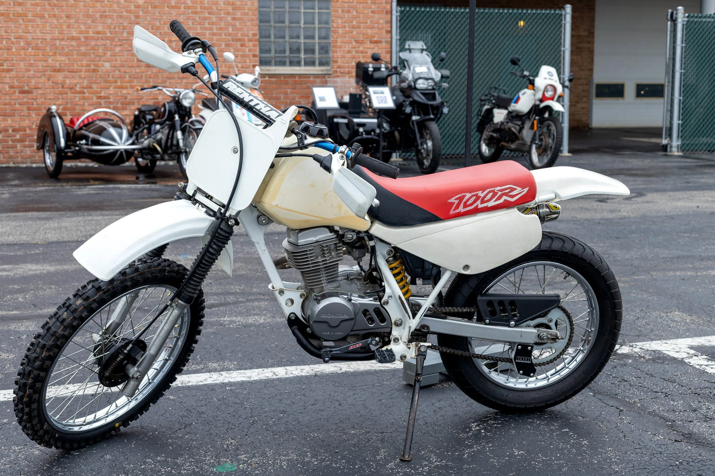 A white off-road motorcycle with a red seat parked on pavement, with several other motorcycles in the background behind a brick wall.