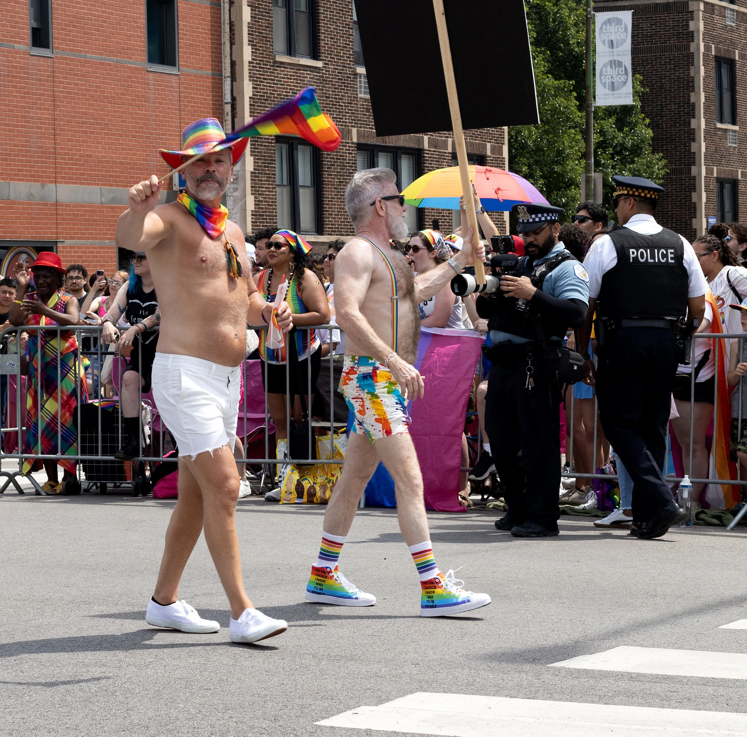 A group of people participating in a pride parade, some are topless and wearing rainbow-colored accessories, with rainbow flags and umbrellas, being watched by police officers. The focus is on two men with rainbow-themed clothing and accessories, inc