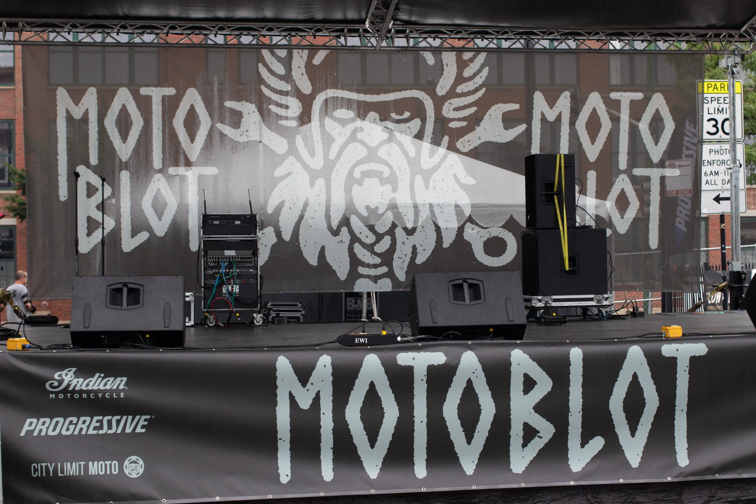 Stage with speakers and audio equipment in front of a backdrop featuring a stylized lion face logo, with text partially visible including 'Indian Motorcycle,' 'Progressive,' and 'City Limit MOTO.'