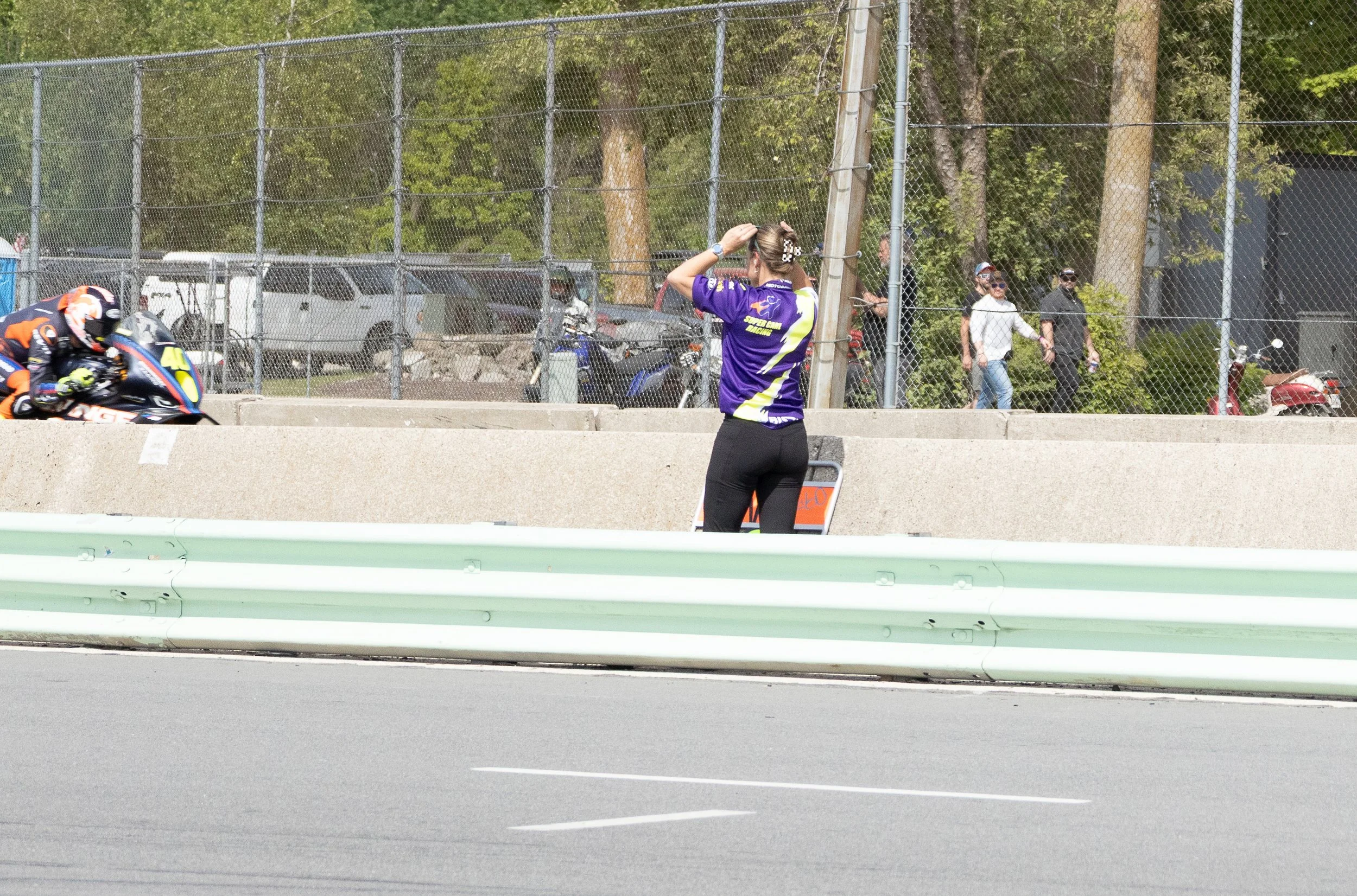 A woman wearing a purple racing shirt stands behind a barrier at a race track, watching a motorcycle racer in orange and black gear lean into a turn. In the background, there are cars, motorcycles, and spectators behind a chain-link fence surrounded 