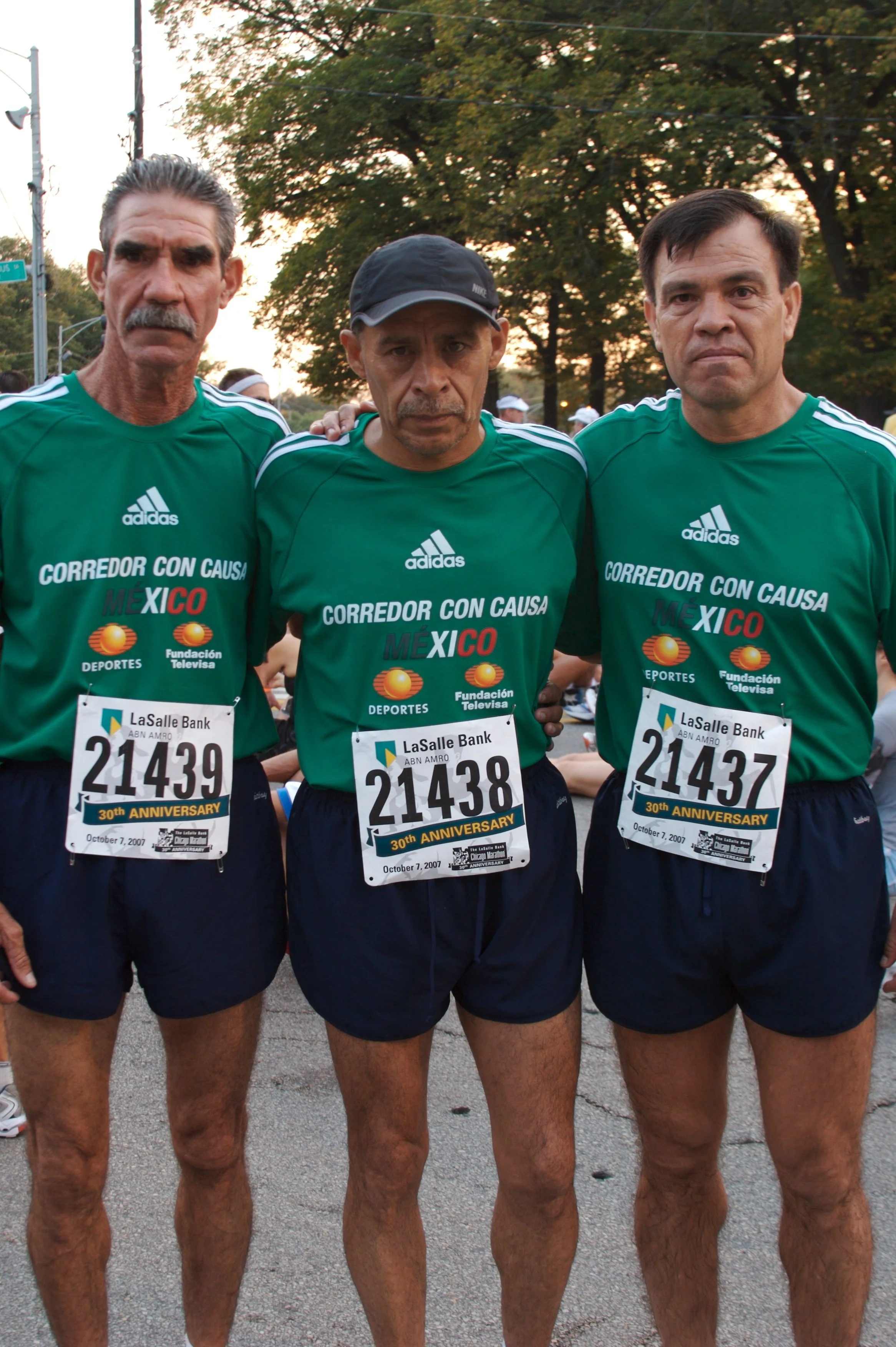 Three men standing side by side wearing green shirts with race bibs, participating in a running event, outdoors with trees and a sunset in the background.