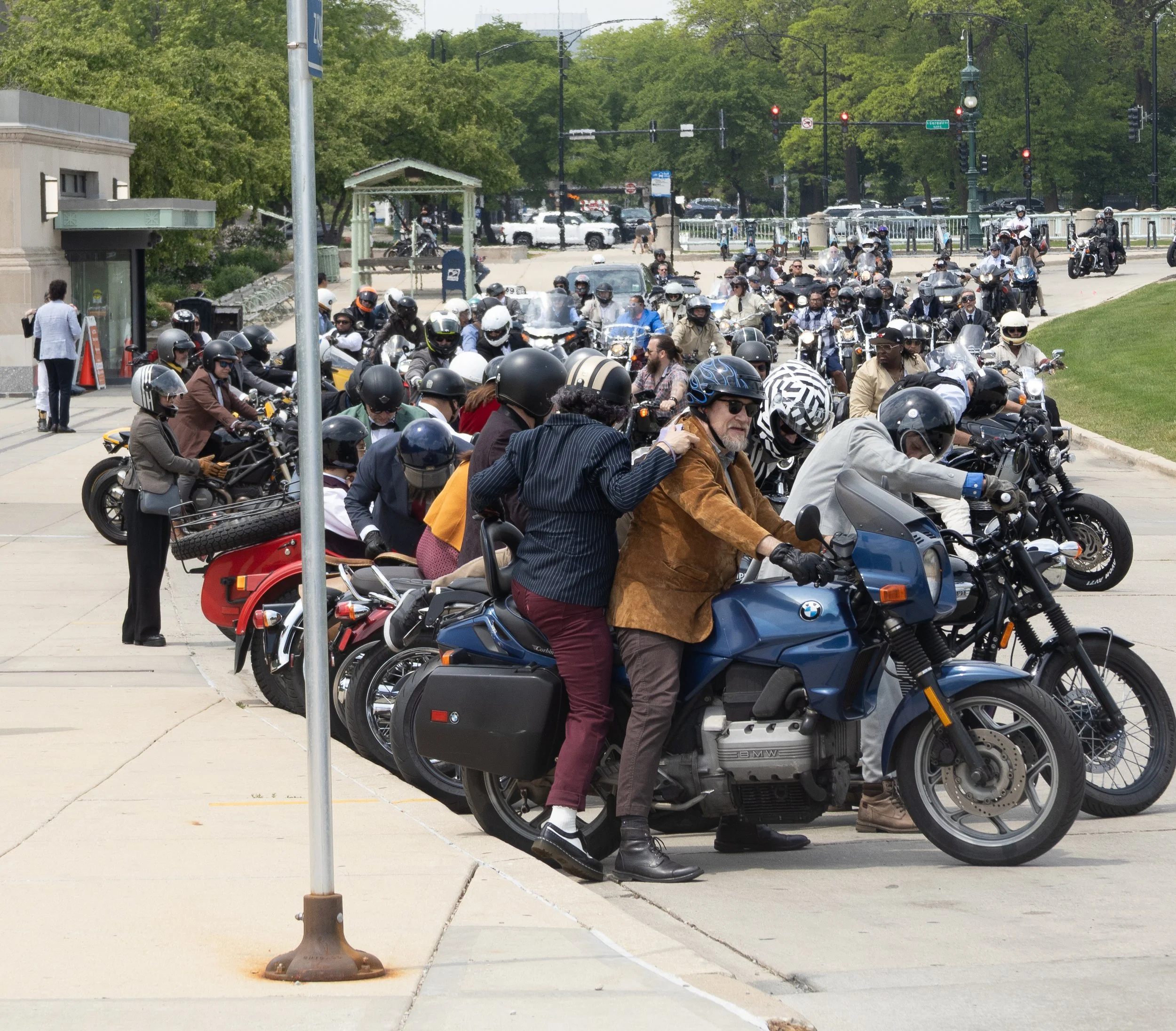A large group of motorcyclists gathered in a parking lot, many wearing helmets and protective gear, with some people walking nearby and a background of trees and traffic signals.
