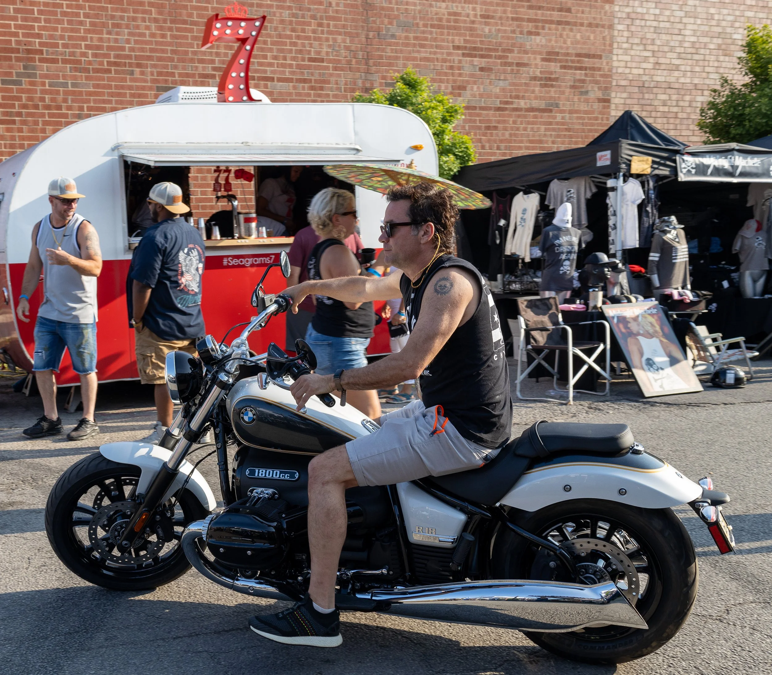 A man with sunglasses and tattoos riding a white BMW motorcycle at an outdoor market or festival. There are people walking and a food and merchandise stand behind him against a brick wall.