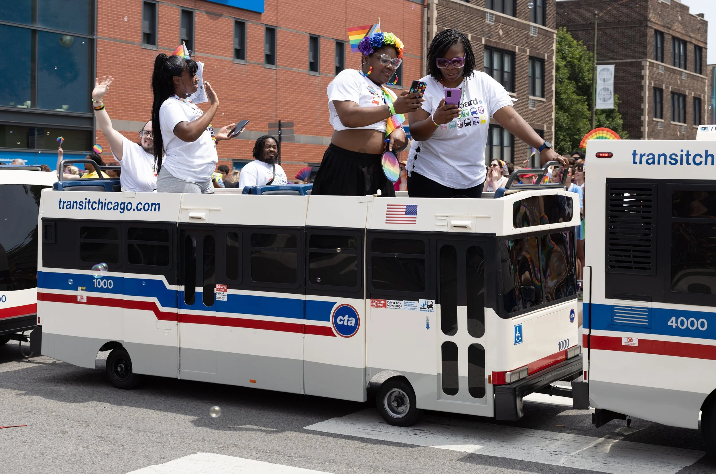 People on a pride parade float decorated with rainbow-themed accessories, looking at their phones and smiling, with a crowd and city buildings in the background.