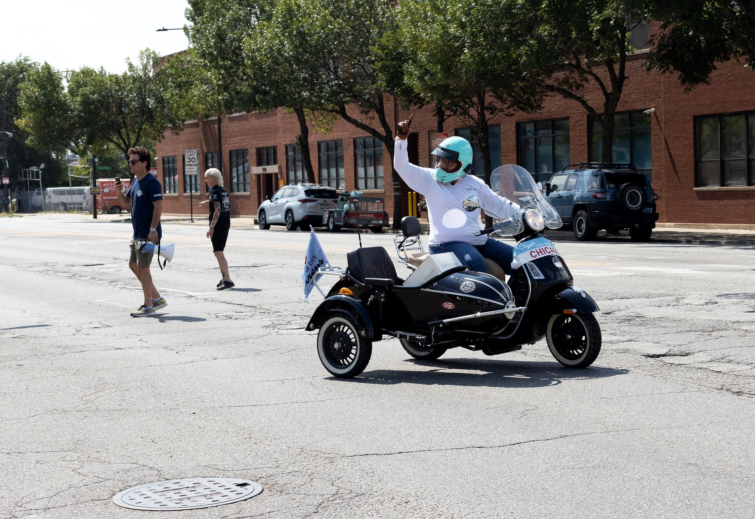 A person riding a motorcycle with a sidecar, wearing a helmet and white shirt, raising one hand with a peace sign during a street event. Pedestrians walk nearby on the crosswalk.