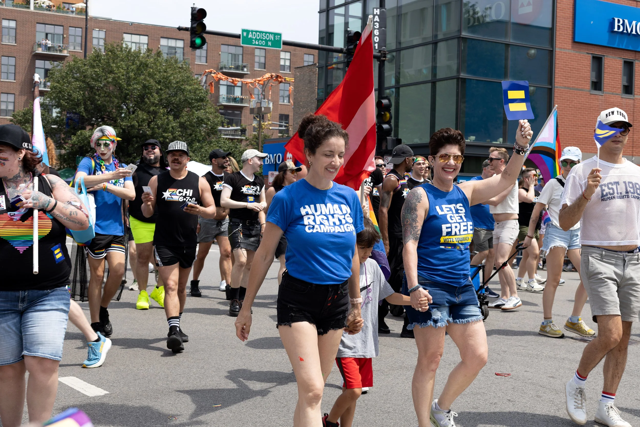 A diverse group of people participating in a pride parade, walking hand in hand, with some holding flags and smiling on a city street with buildings and green traffic lights in the background.