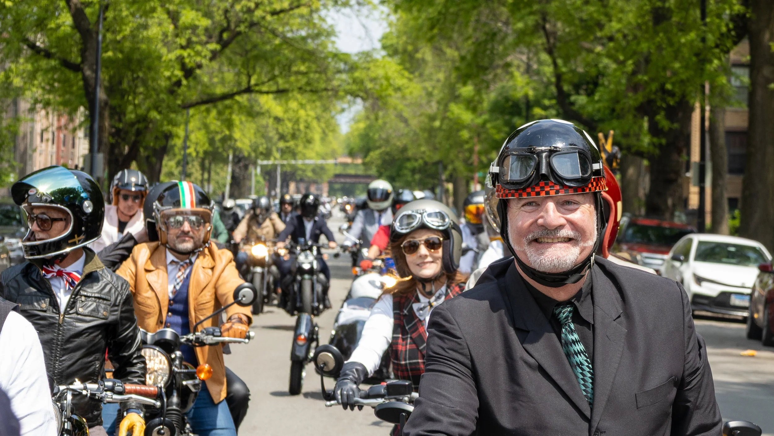 A group of people riding motorcycles on a city street, wearing helmets and sunglasses, with green trees and parked cars in the background.