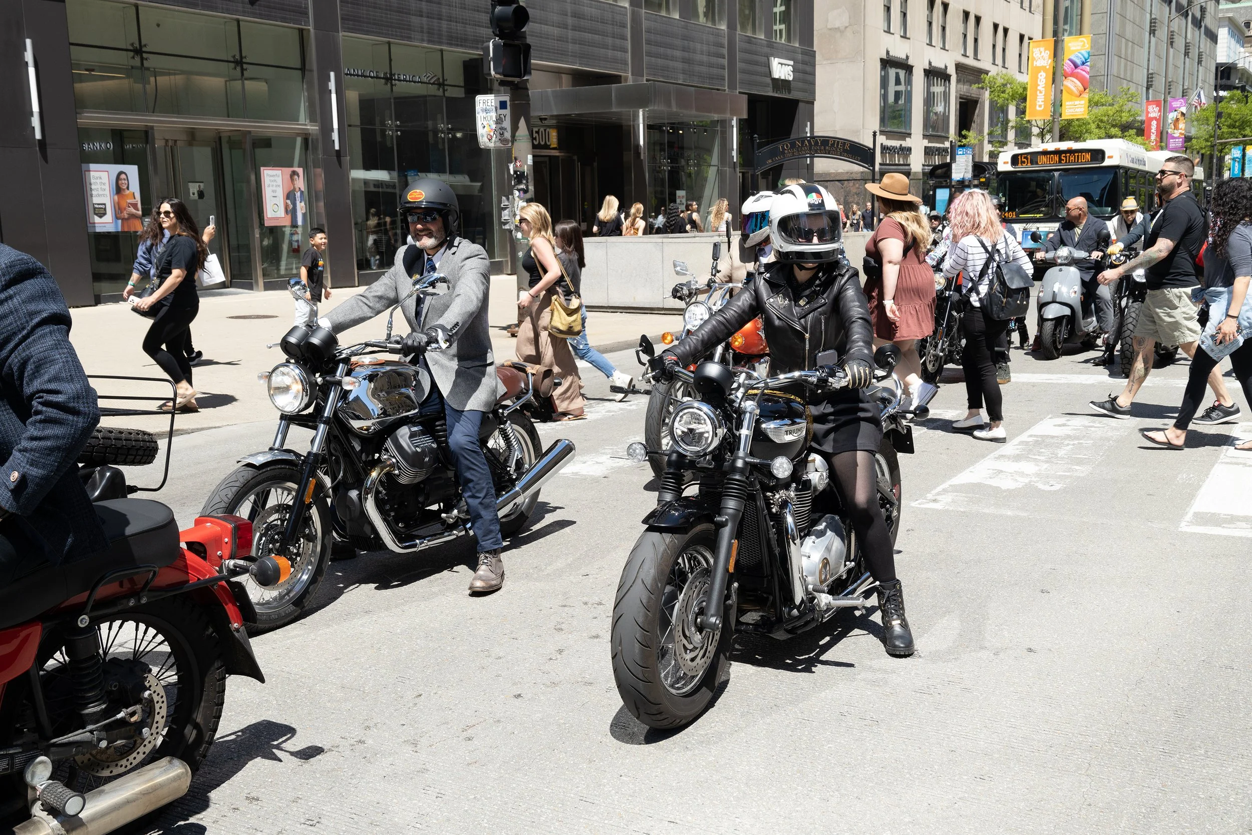 Crowded city street with people walking and riding motorcycles, storefronts, and bus on a sunny day.