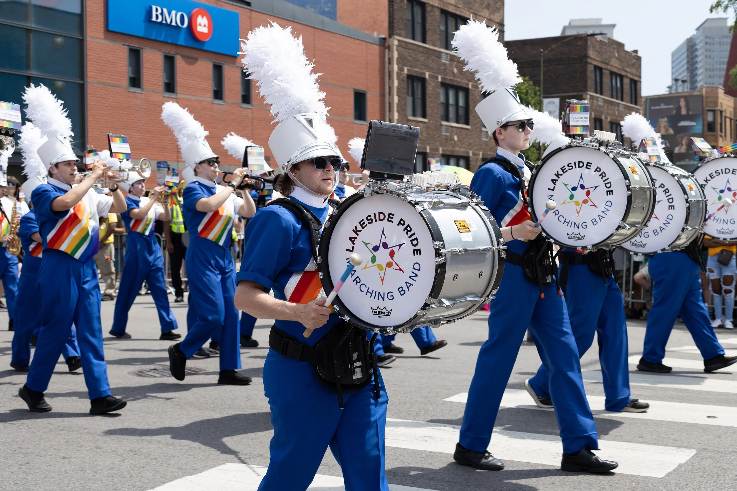 A marching band performing in a parade, with members dressed in blue uniforms and white hats with large feather plumes, playing drums labeled 'Lakeside Pride Marching Band,' in an urban area with buildings and spectators in the background.
