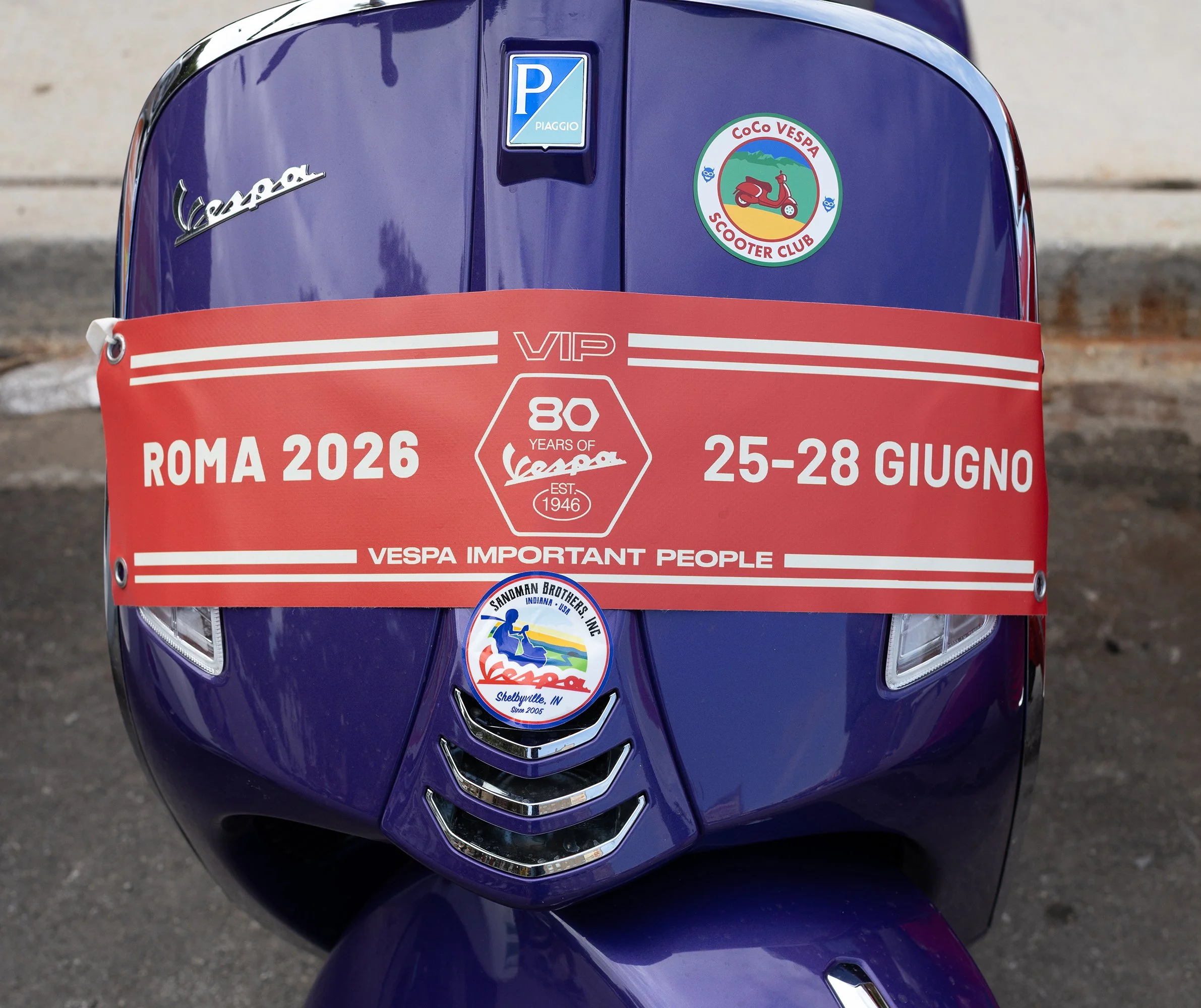 The front of a blue Vespa scooter with various stickers, including a Vespa logo, a parking sticker, and a round club sticker featuring a Vespa scooter, mountains, and a sunset.