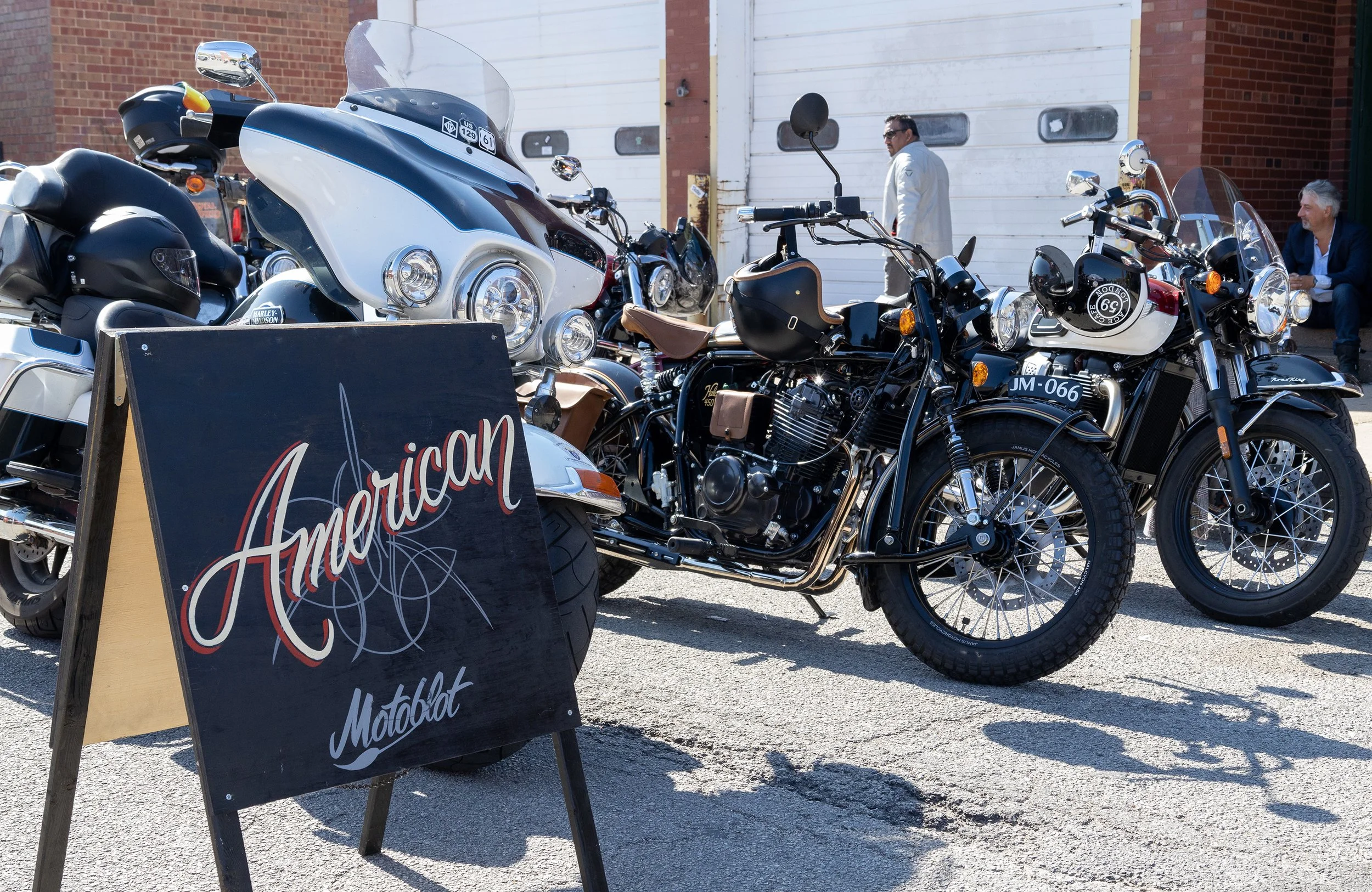 A lineup of motorcycles parked outdoors in front of a white garage door, with a restaurant-style blackboard sign reading 'American' in front, and two men in the background talking, one sitting and one standing near the bikes.