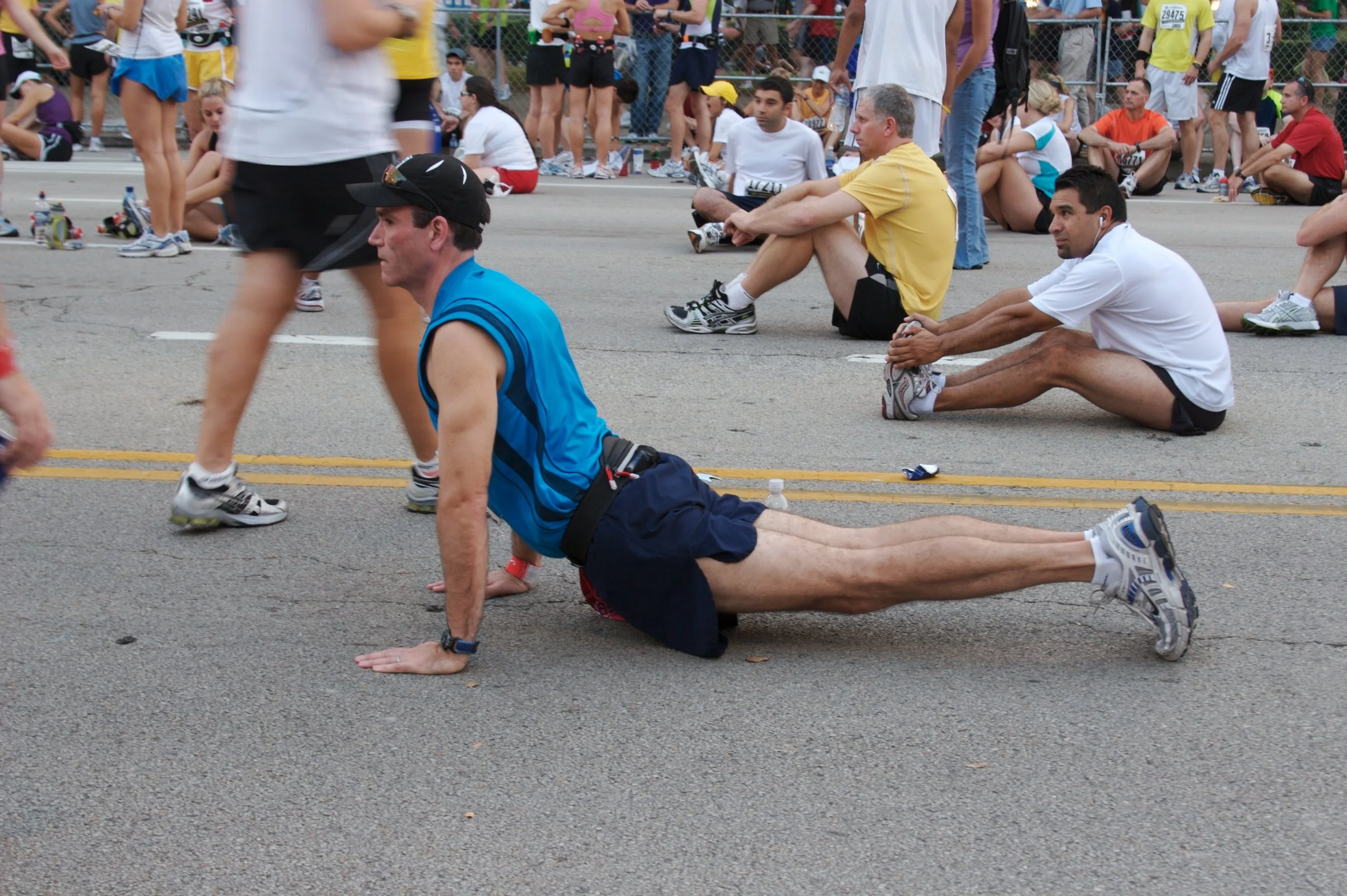 People participating in a race; some are taking a break and sitting on the ground, others are walking or stretching on a paved street with a crowd behind a fence.