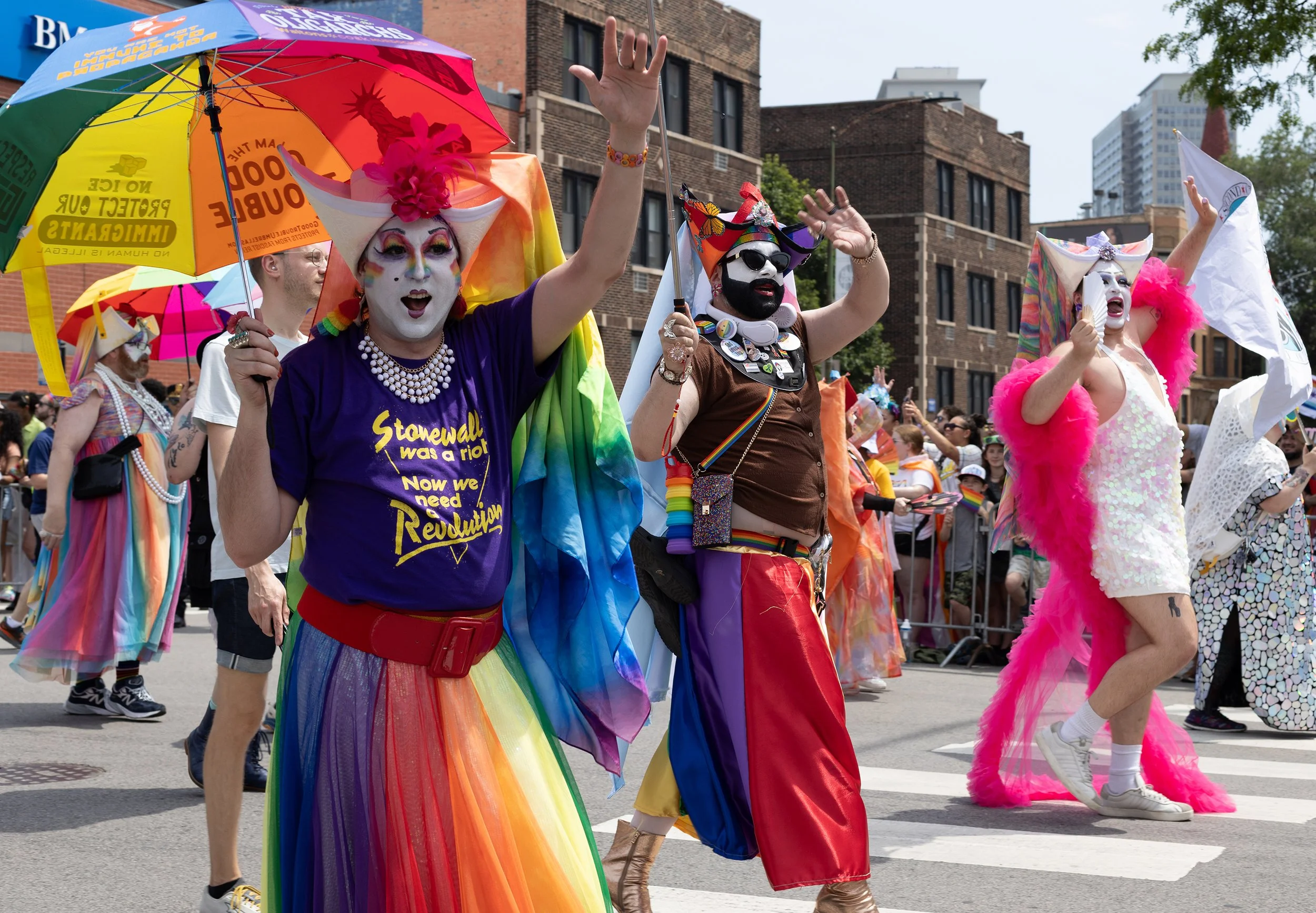 People dressed in colorful rainbow costumes participating in a pride parade. They are waving and holding umbrellas, with cheerful expressions and vibrant makeup.