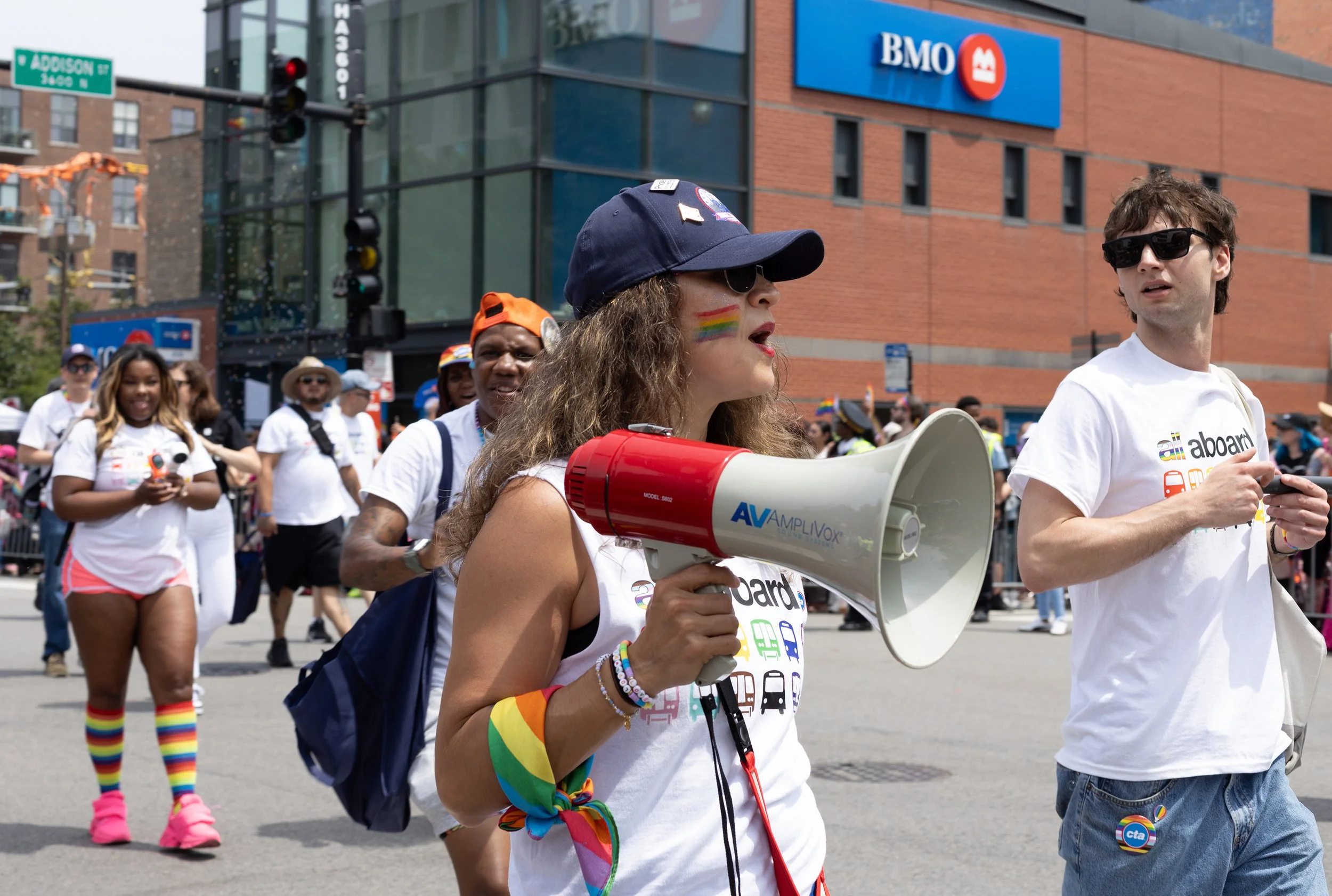 A group of people participating in a pride parade in front of a BMO bank building, some wearing rainbow-themed clothing and accessories. A woman in the foreground is speaking into a megaphone, wearing rainbow wristbands and face paint. The crowd appe