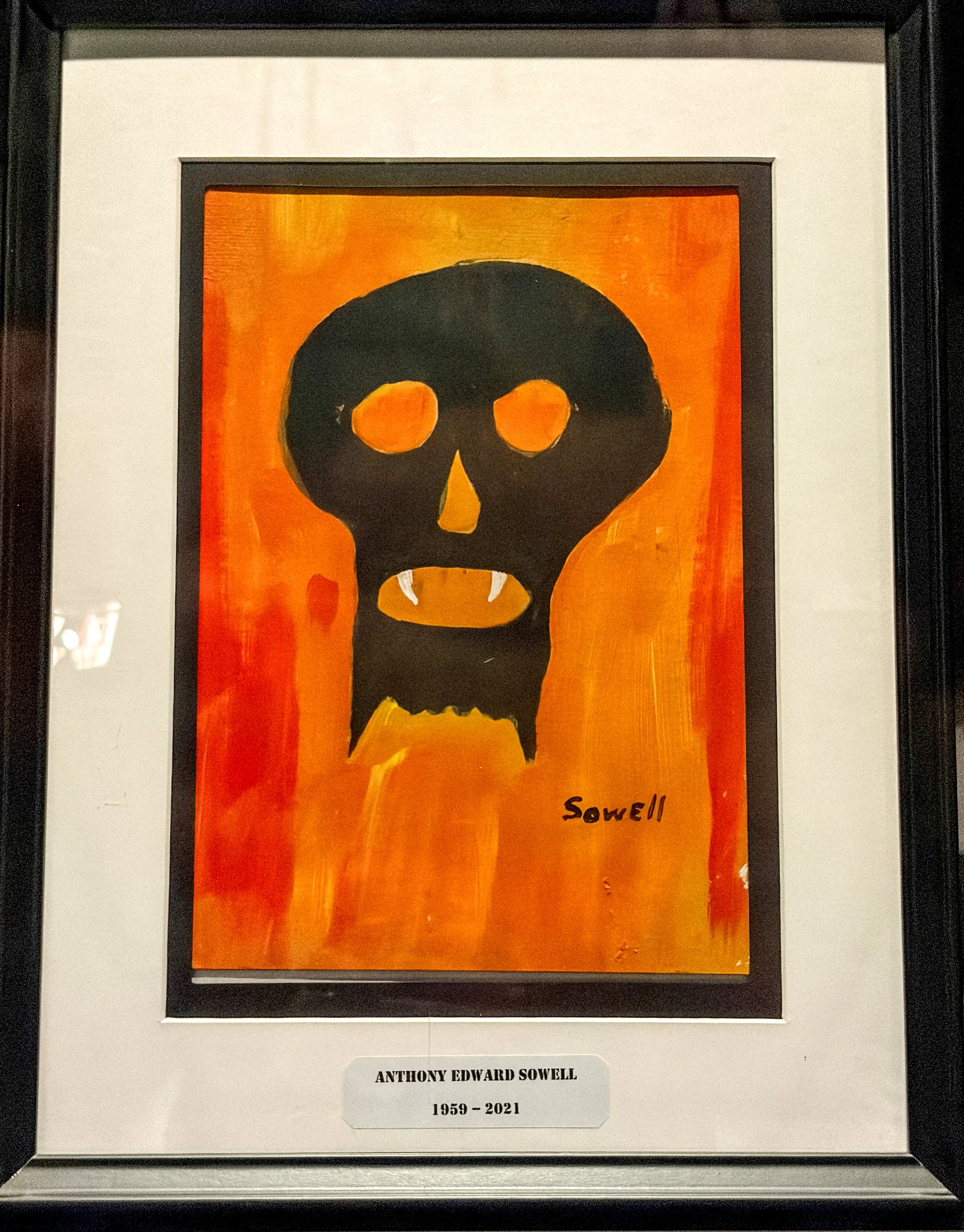 Framed artwork of a black skull on a bright orange background. The artwork is signed as 'Sowell' and has a plaque below it with the name 'Anthony Edward Sowell' and the years '1959-2021'.