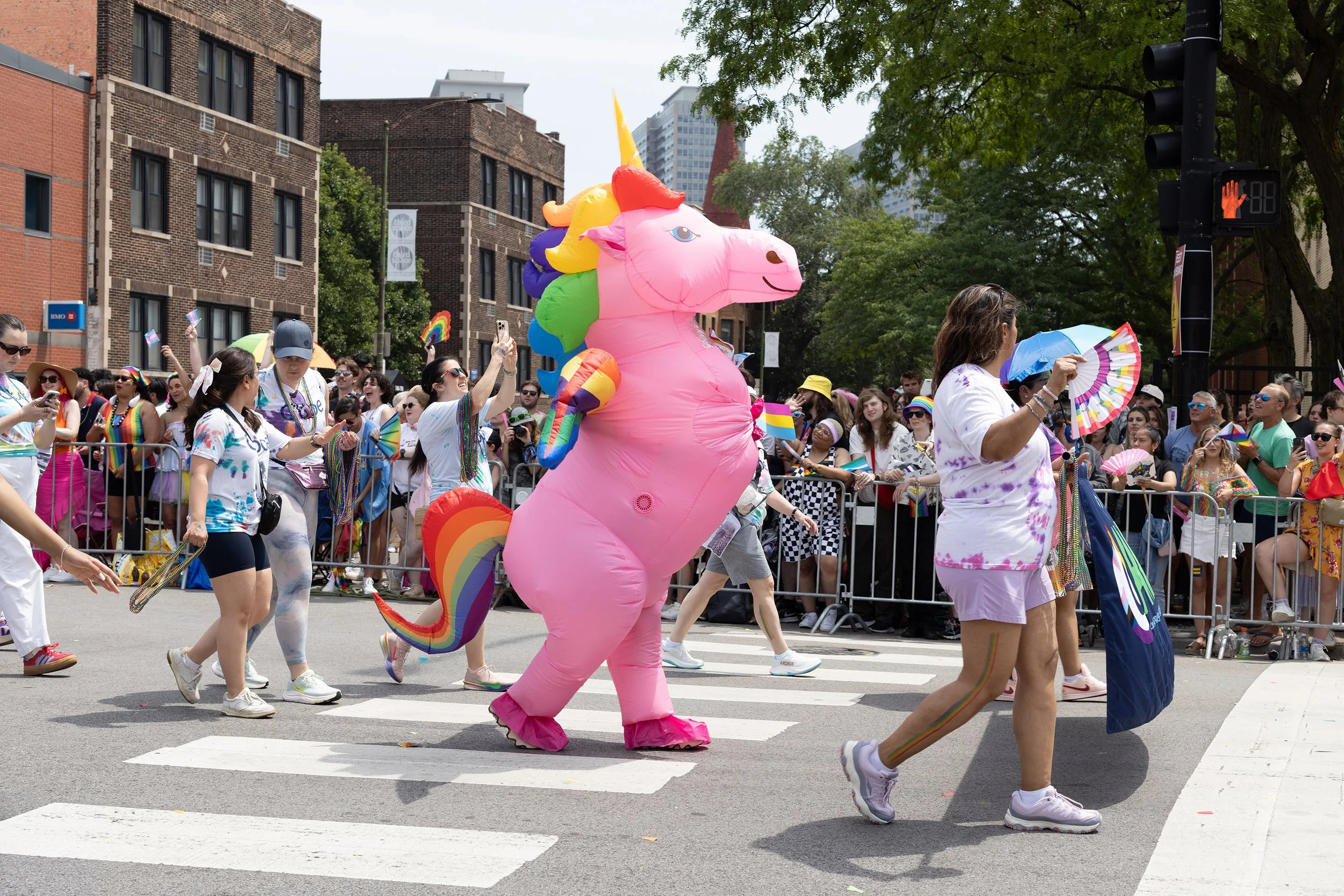 People participating in a pride parade on a city street, with a large inflatable pink unicorn in the center, and many spectators behind a barrier, some waving rainbow flags, wearing colorful accessories, with city buildings and trees in the backgroun