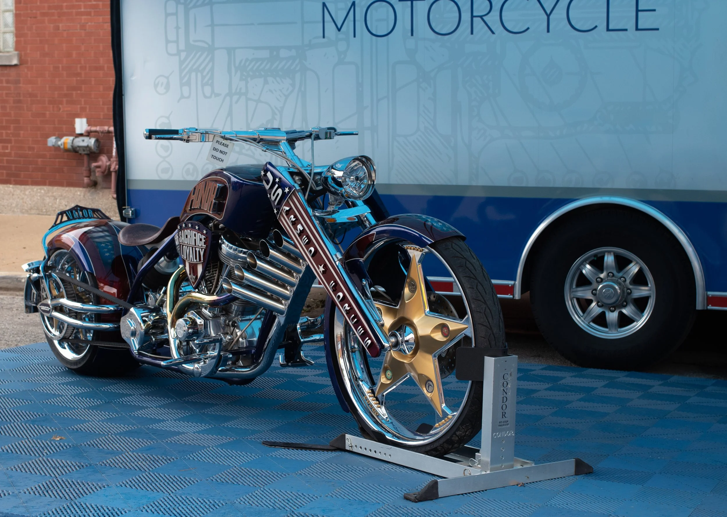 A shiny, custom motorcycle on display outdoors, with a sign reading 'Please Do Not Touch,' and a trailer in the background with the word 'Motorcycle' partially visible.