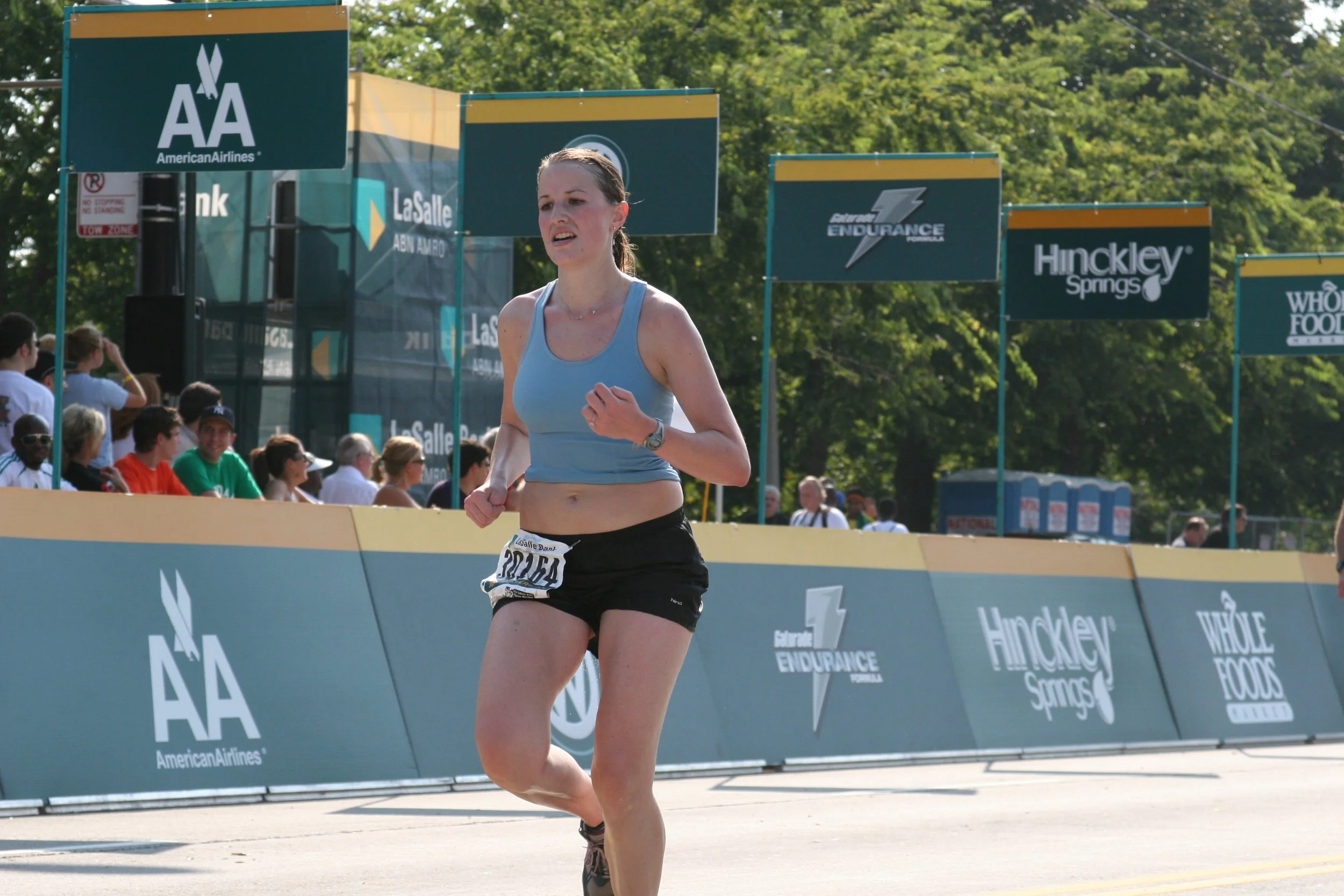 A female runner in athletic clothing, running in a race event with spectators and advertising banners in the background.