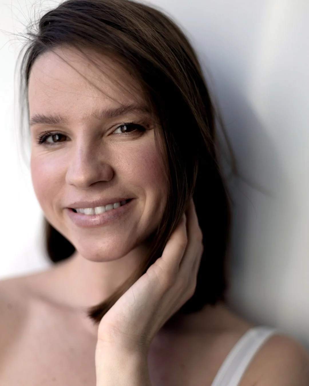 A close-up portrait of a woman with brown hair, smiling, with her hand gently touching her neck, against a plain background.