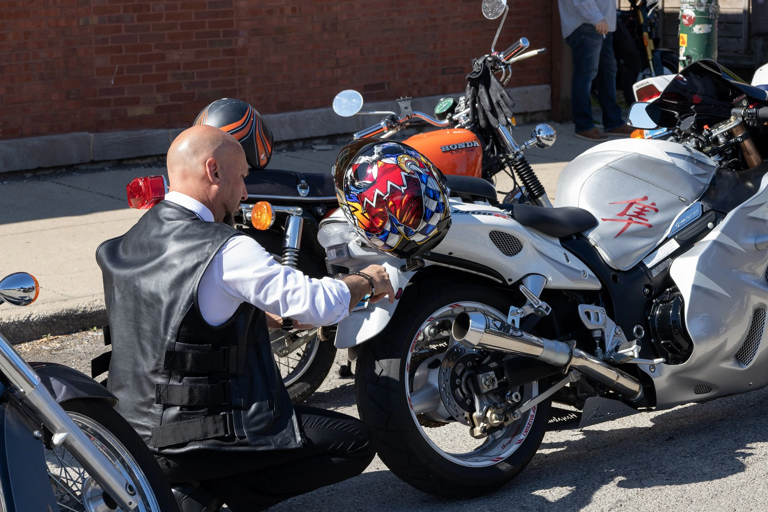 A man in a white shirt and black leather vest sitting on a white Harley-Davidson motorcycle, with a colorful helmet on the handlebar, parked alongside an orange Honda motorcycle with a black and orange helmet on top. In the background, a brick wall a