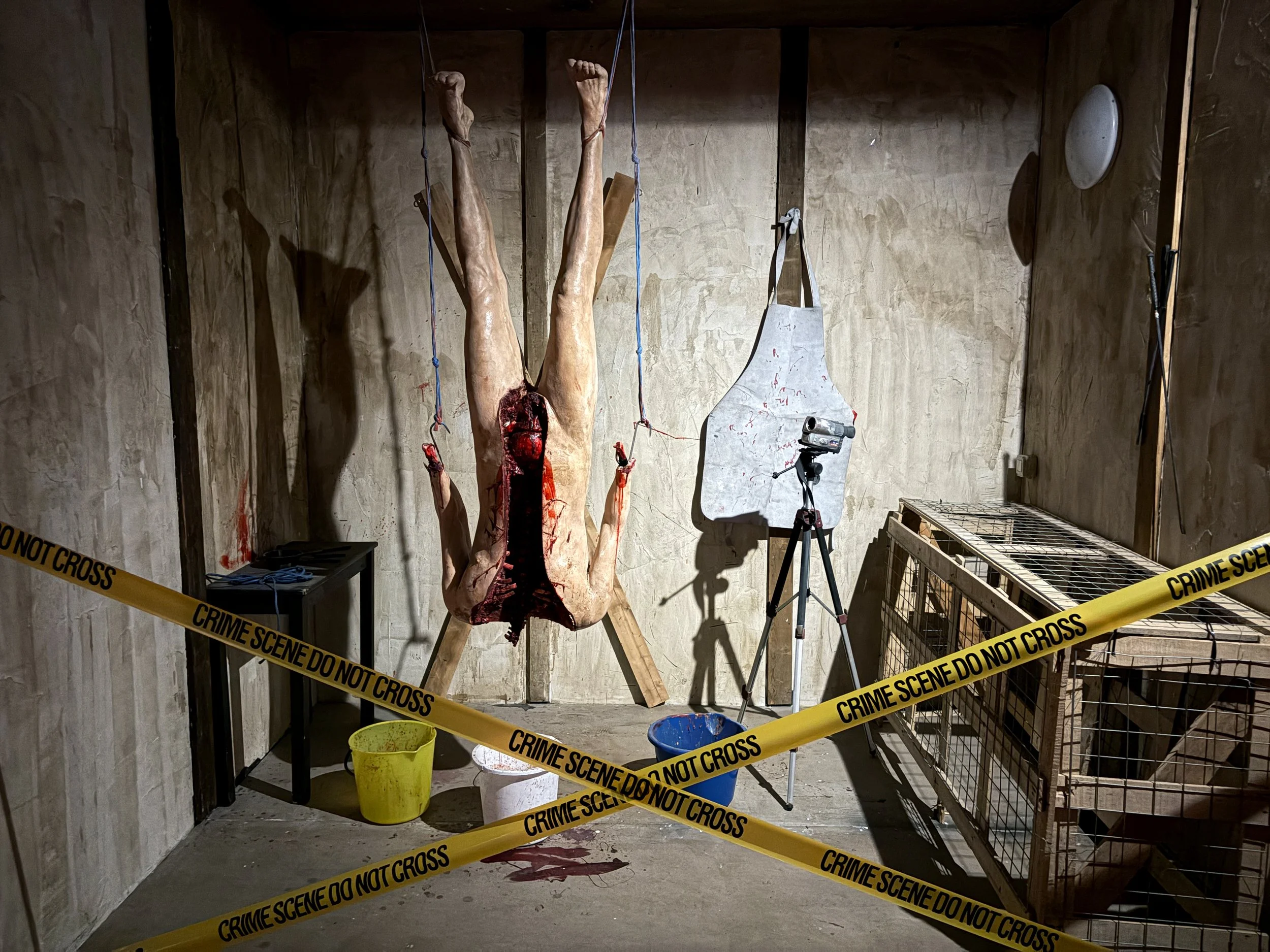 A realistic-looking sculpture of a severed human leg and foot, with exposed muscle tissue, hanging upside down from the ceiling, surrounded by yellow crime scene tape, and warning signs, in an unfinished room with wooden walls and a concrete floor.