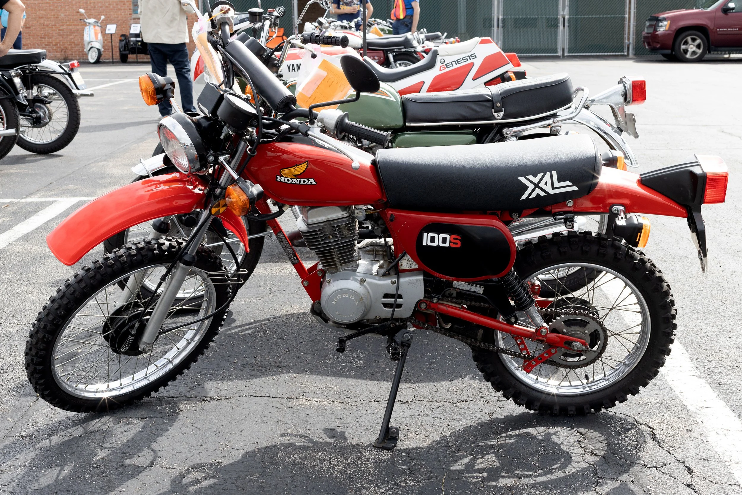 A red Honda dirt bike with knobby tires parked in a lot, with additional motorcycles and people in the background.