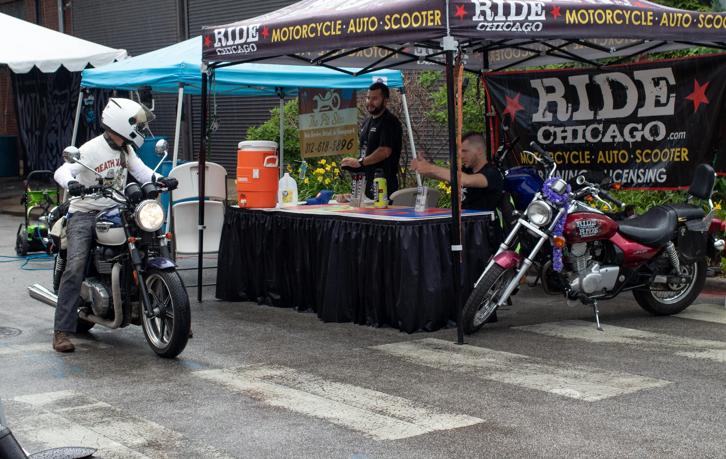 A motorcycle rider wearing a white helmet and a white long-sleeve shirt, sitting on a motorcycle at a booth with a banner that reads 'RIDE CHICAGO MOTORCYCLE AUTO SCOOTER.' The booth has a table with supplies, and two men are standing behind it, one 