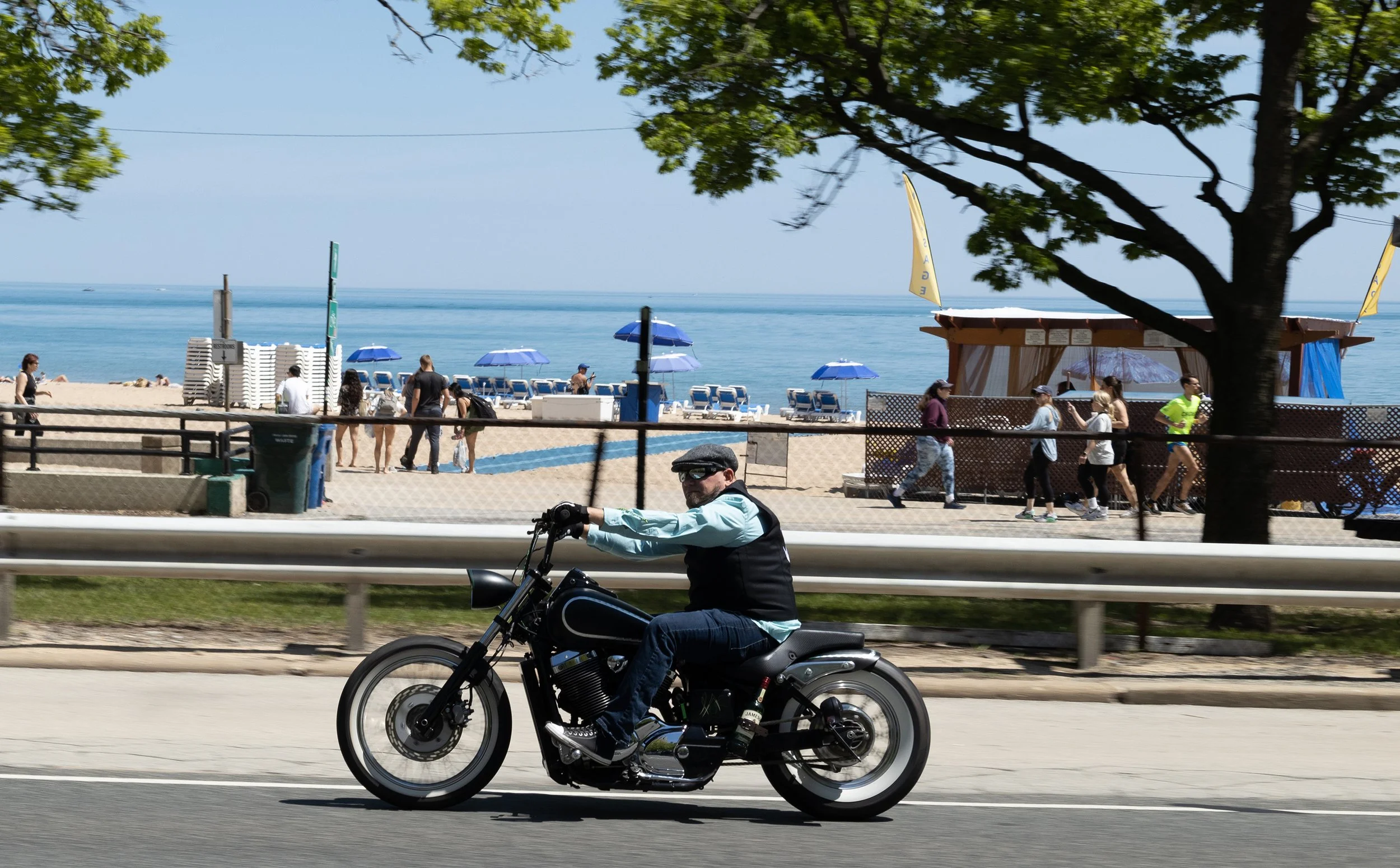 A man riding a black motorcycle along a road near a beach with beach chairs and umbrellas, a large tree, and people walking near the beach.
