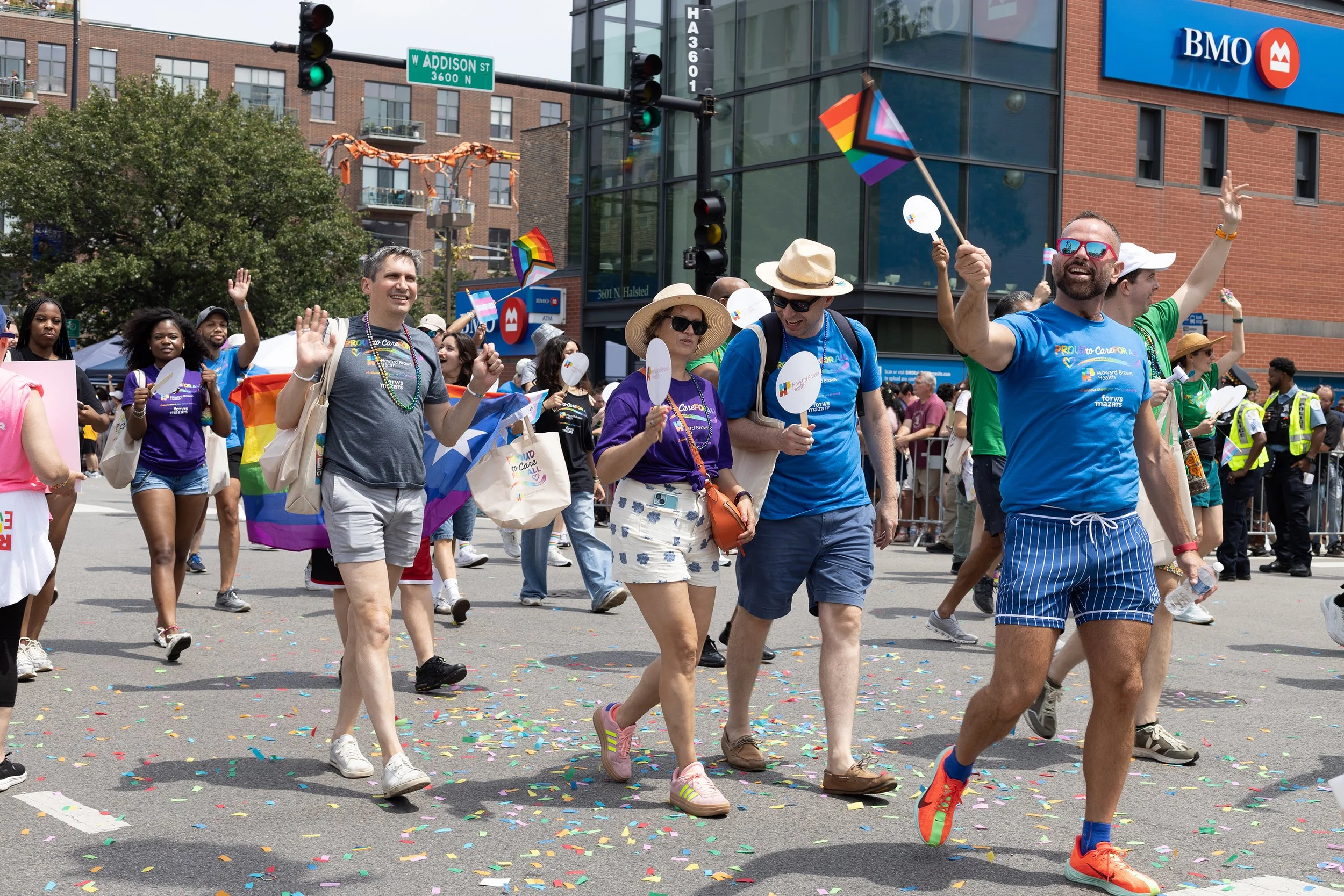 A group of people celebrating in a pride parade, walking on a street with colorful confetti on the ground, holding pride flags, and wearing casual summer clothing.