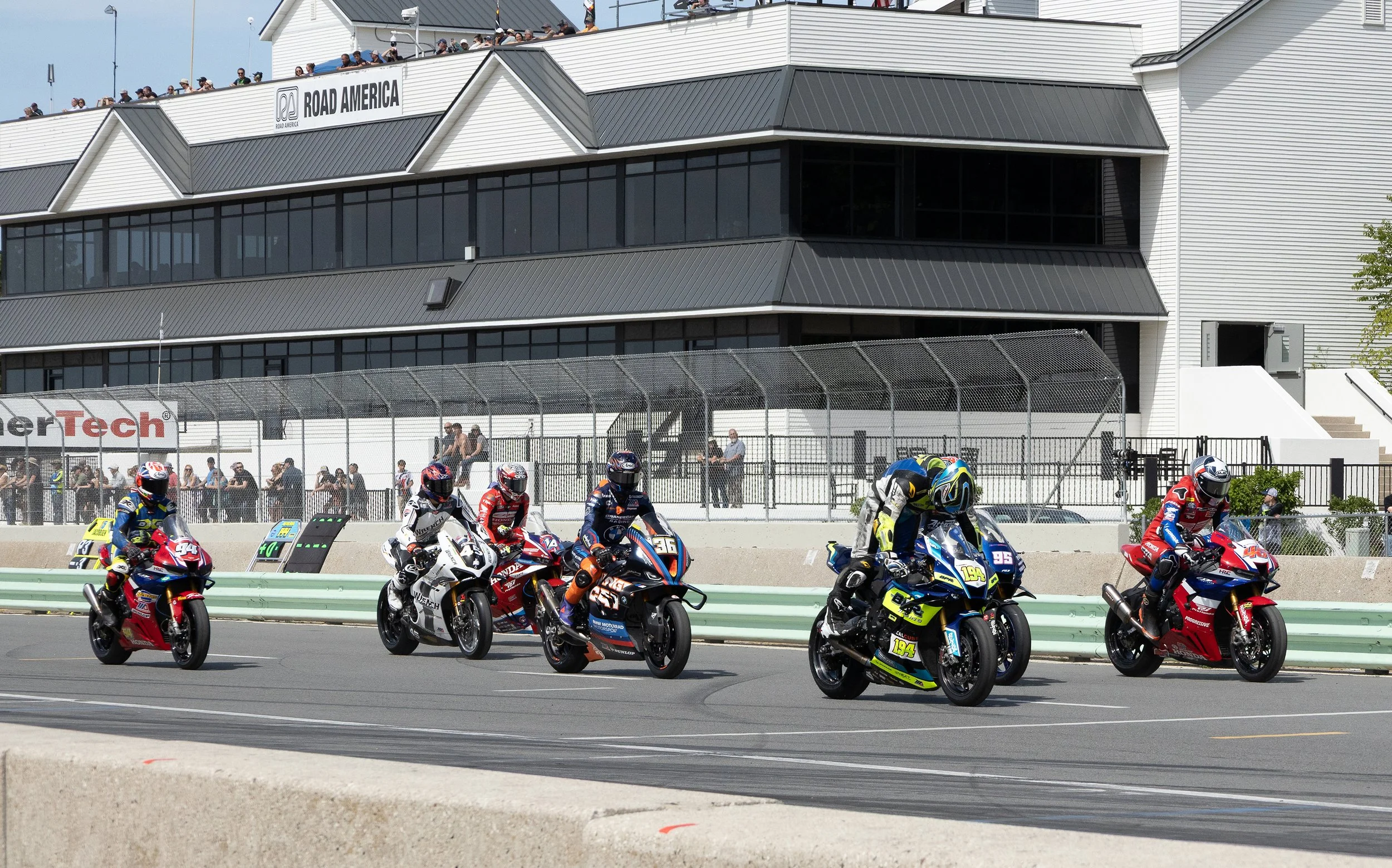 Motorcycle racers lined up at the start of a race on the track, with spectators watching from the grandstand and roof of the building behind them.