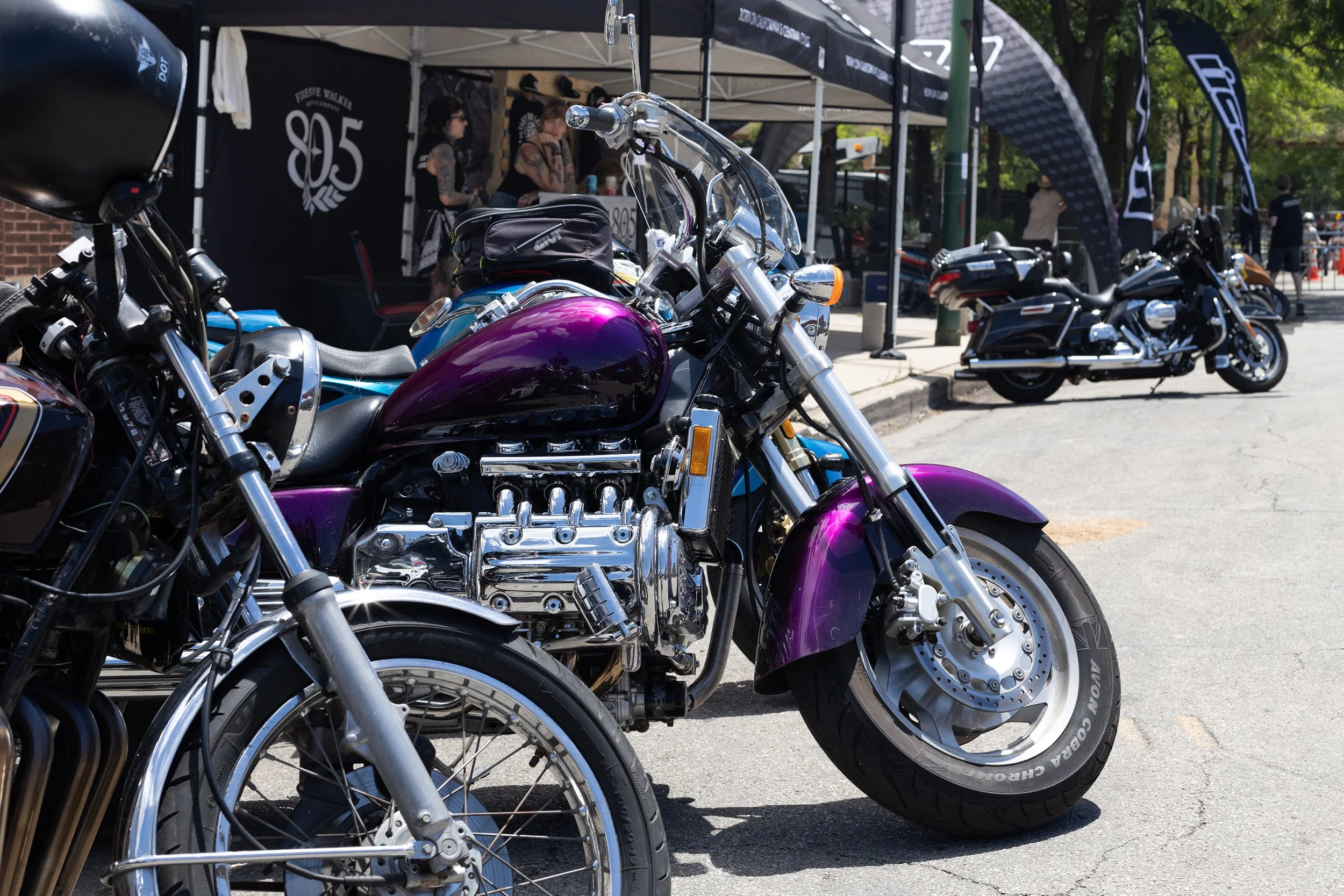 Several motorcycles parked in front of a booth at an outdoor event with people in the background.