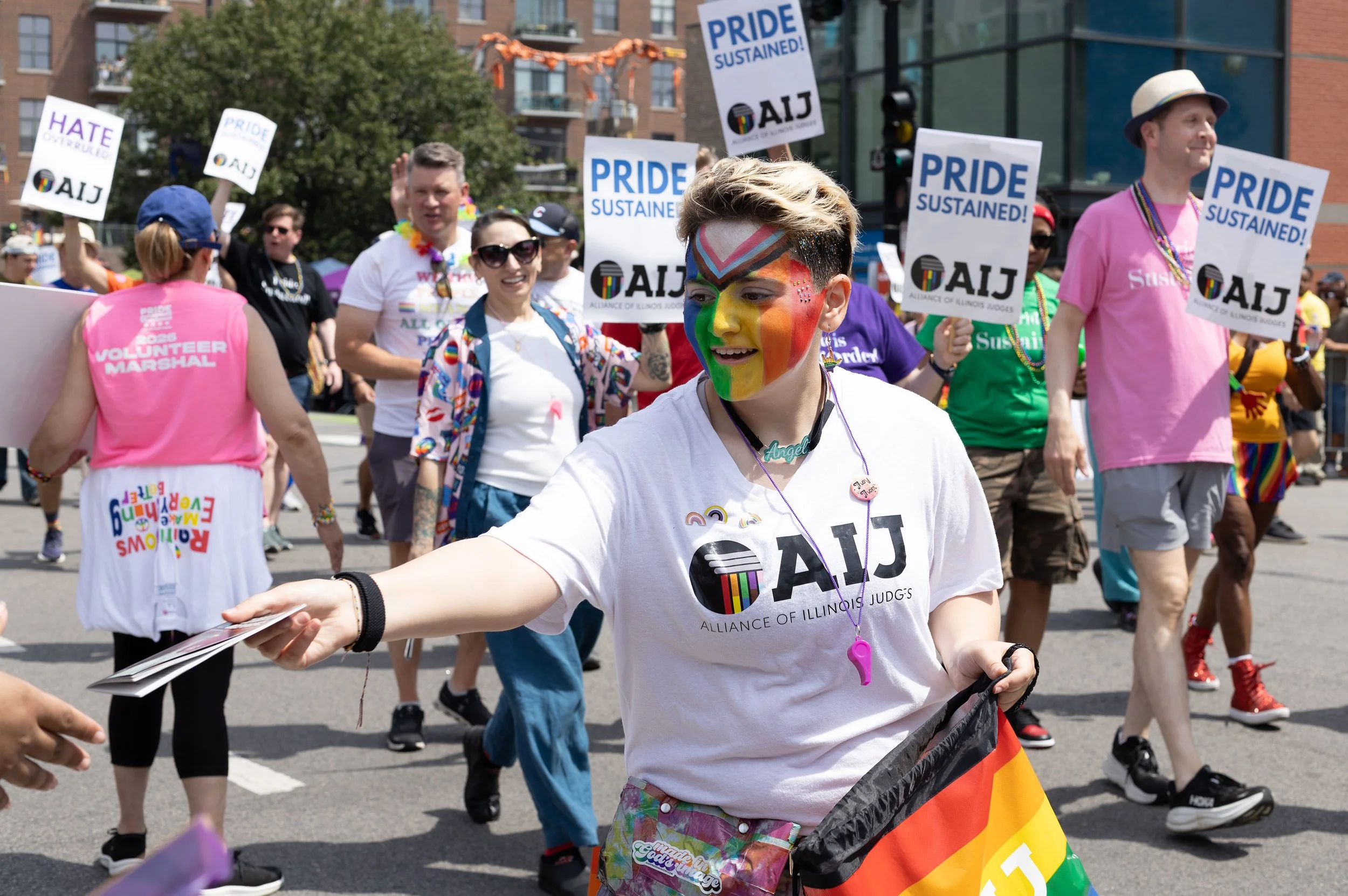 Participants in a Pride parade, holding signs that read 'PRIDE SUSTAINED!' and wearing colorful clothing and accessories. One person in the foreground has rainbow face paint and a necklace that says 'Angel,' and is handing out flyers.