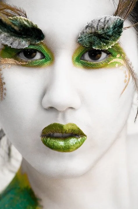 Close-up of a person with dramatic green, gold, and blue makeup, including feathered eyelash extensions and metallic lips.