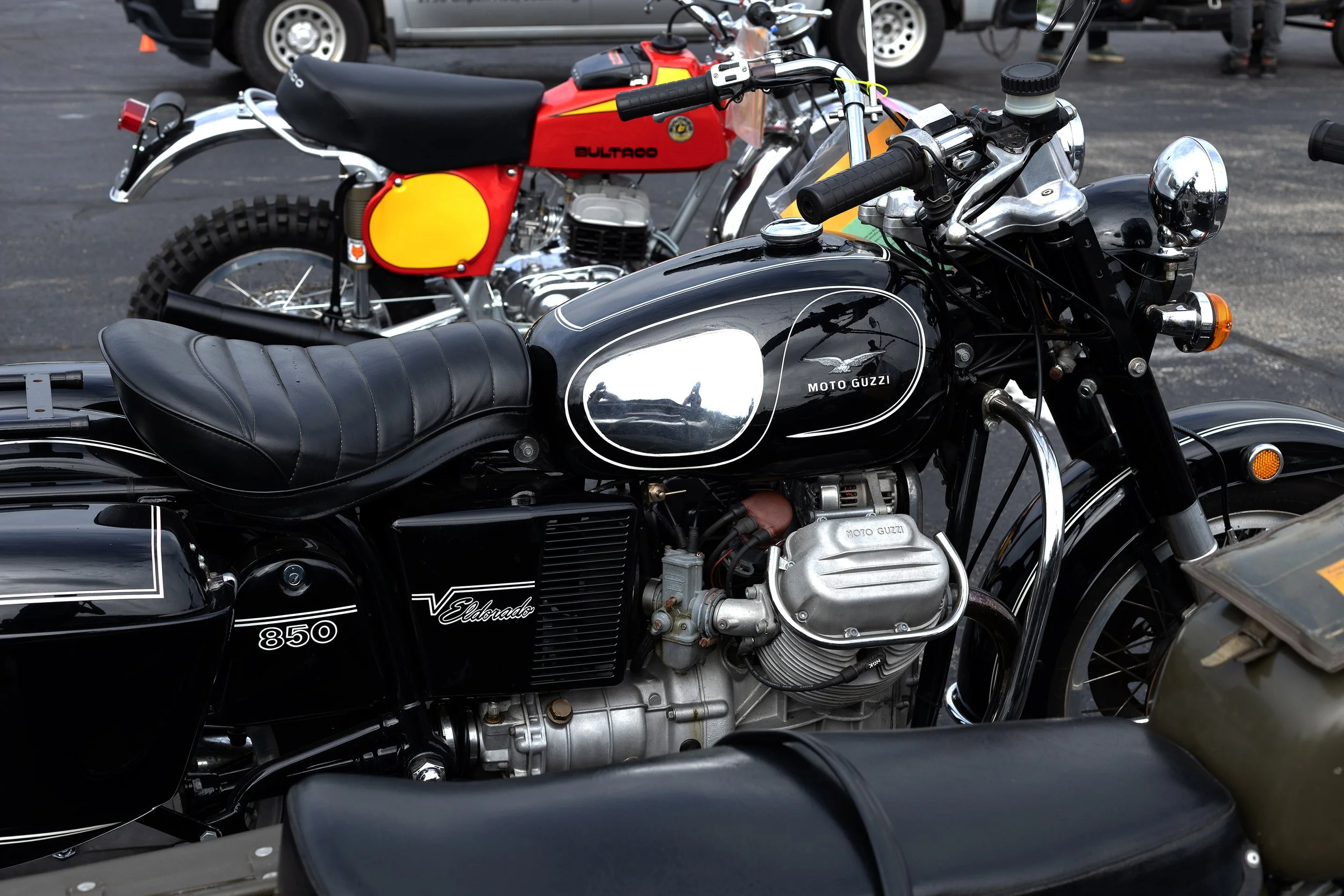 Close-up of vintage motorcycles parked outdoors, including a black Moto Guzzi with chrome accents and a red Bultaco with yellow details in the background.