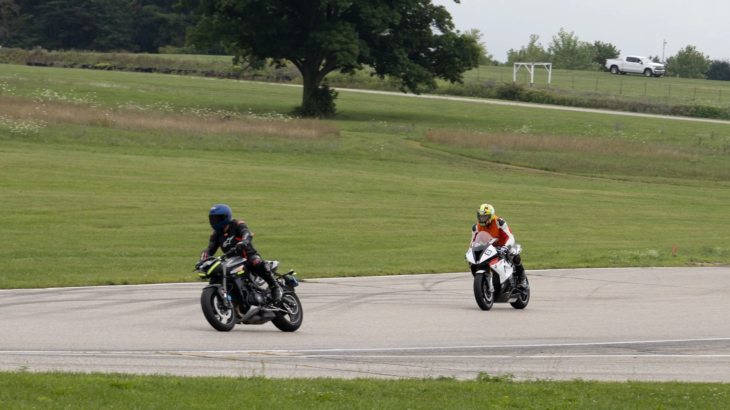 Two motorcyclists riding on a track with a grassy field and trees in the background. One rider is in a black suit with a blue helmet, the other in a white and red suit with a matching helmet.