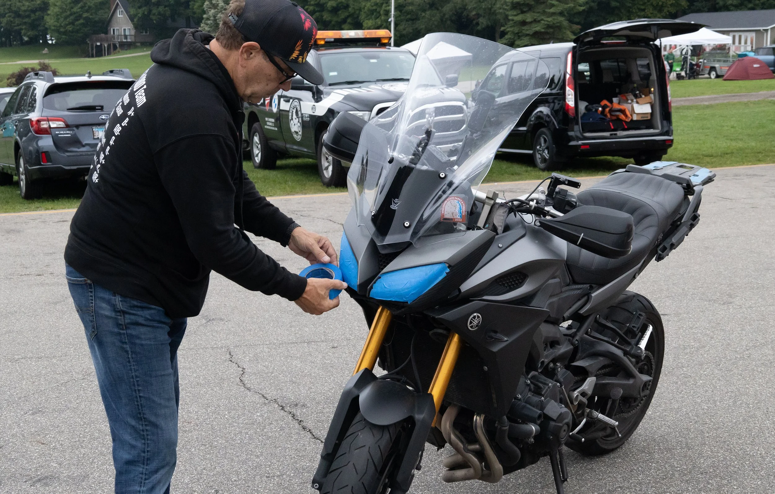 A man wearing glasses, a black hoodie, a black cap with colorful patterns, is fixing or inspecting a Yamaha motorcycle. The motorcycle is parked on the side of a road, with a storage box or container open behind it. Several parked cars and a grassy a