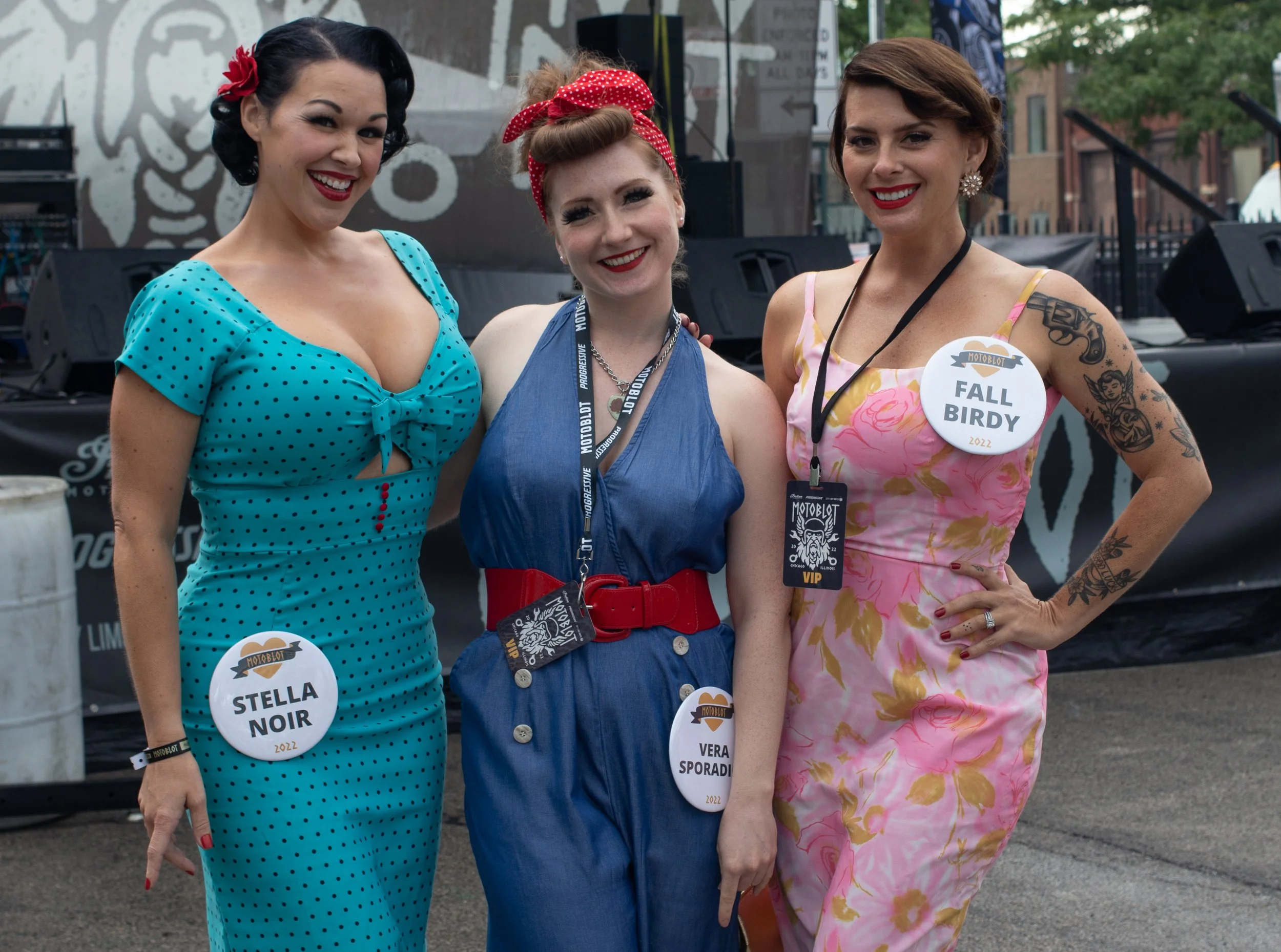 Three women posing at an outdoor event, wearing vintage-style dresses with name tags. The woman on the left has dark hair styled in victory rolls, a red flower hair accessory, and a blue polka-dot dress. The woman in the middle has red hair styled in