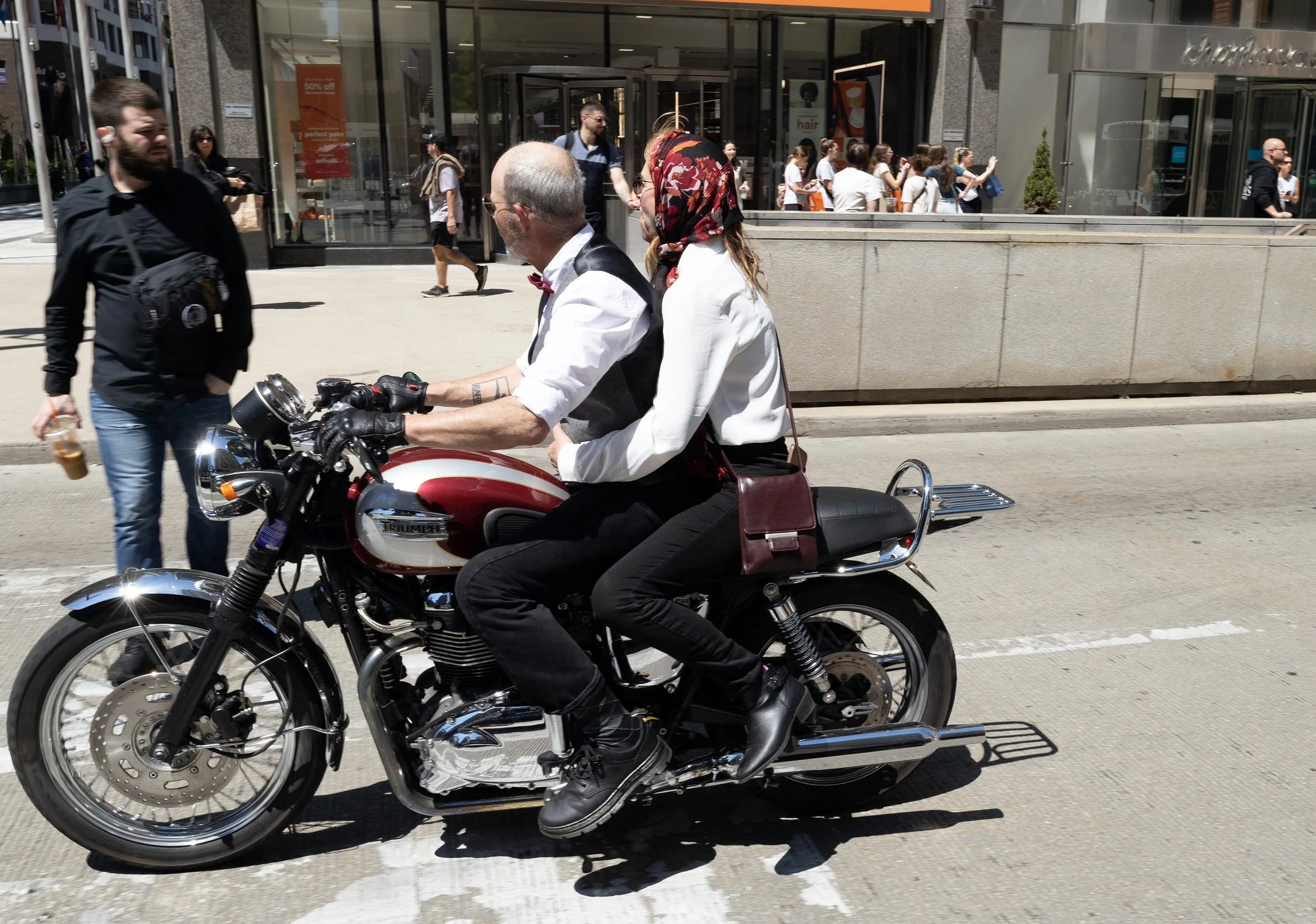 People walking and a man riding a motorcycle on a city street in front of a glass storefront.